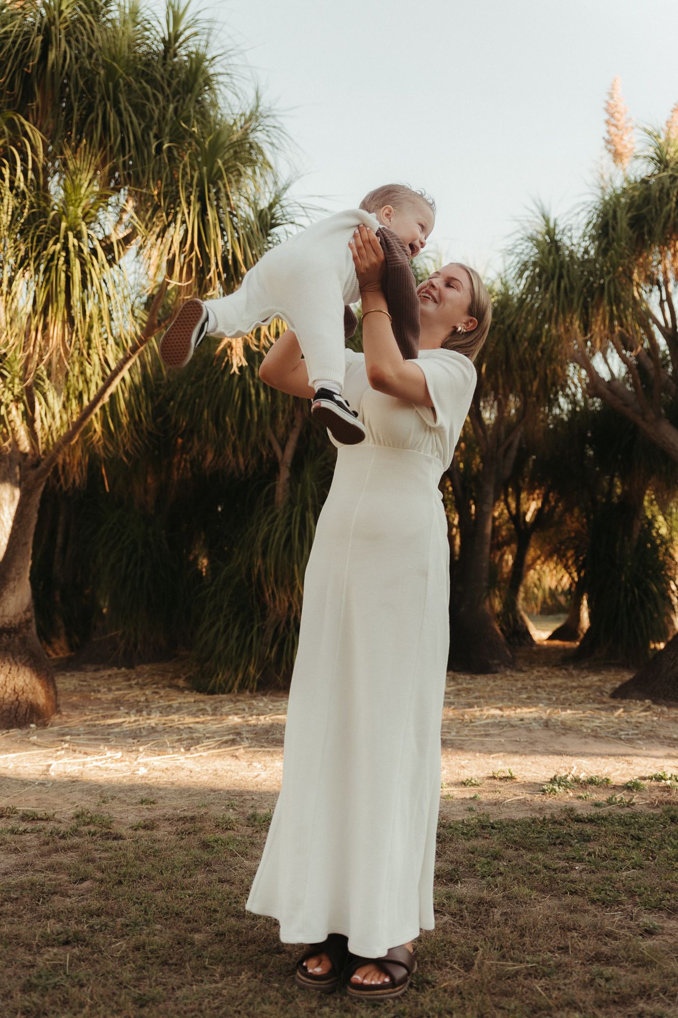 Woman in white dress joyfully lifting a baby outdoors near palm trees in warm sunlight.