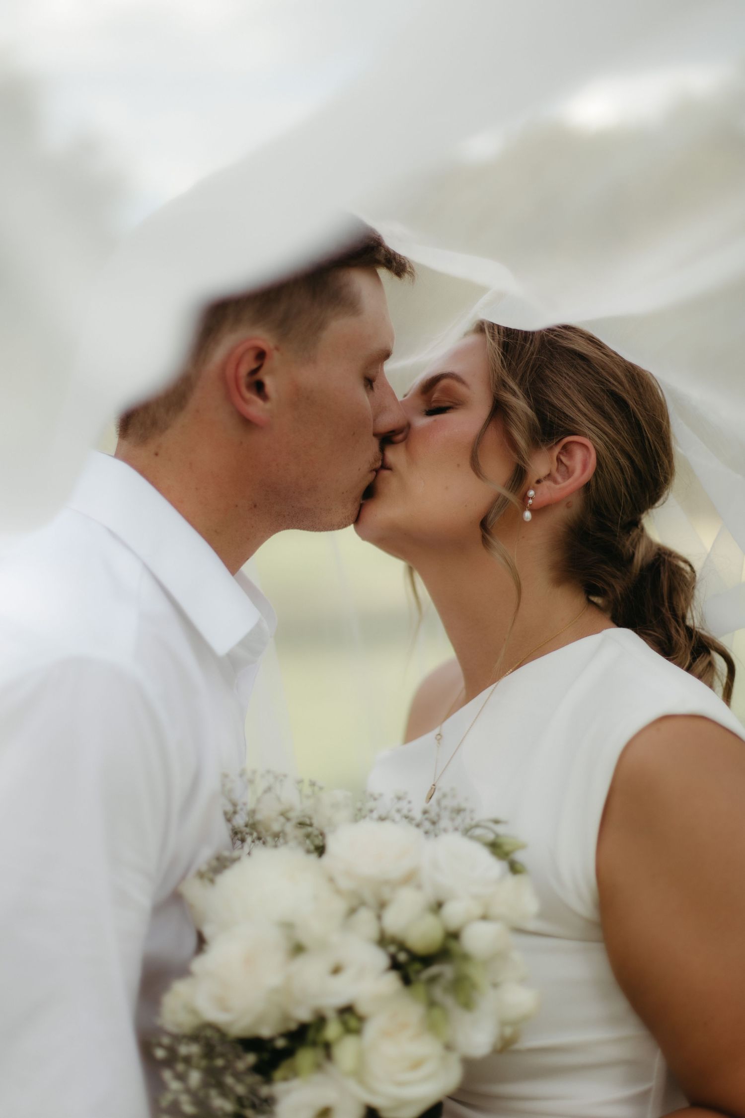 Bride and groom sharing romantic kiss under flowing veil, bride holding white rose bouquet on wedding day.