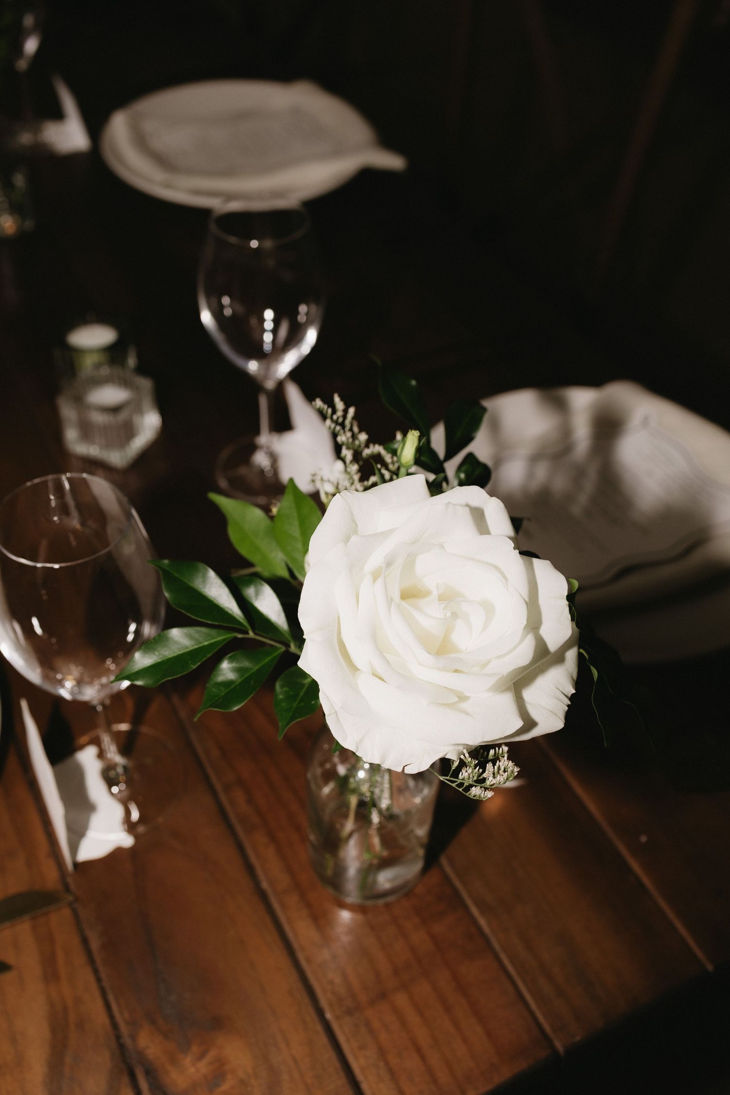 White rose centerpiece in glass vase on wooden table with wine glasses and linen napkins at elegant wedding reception.