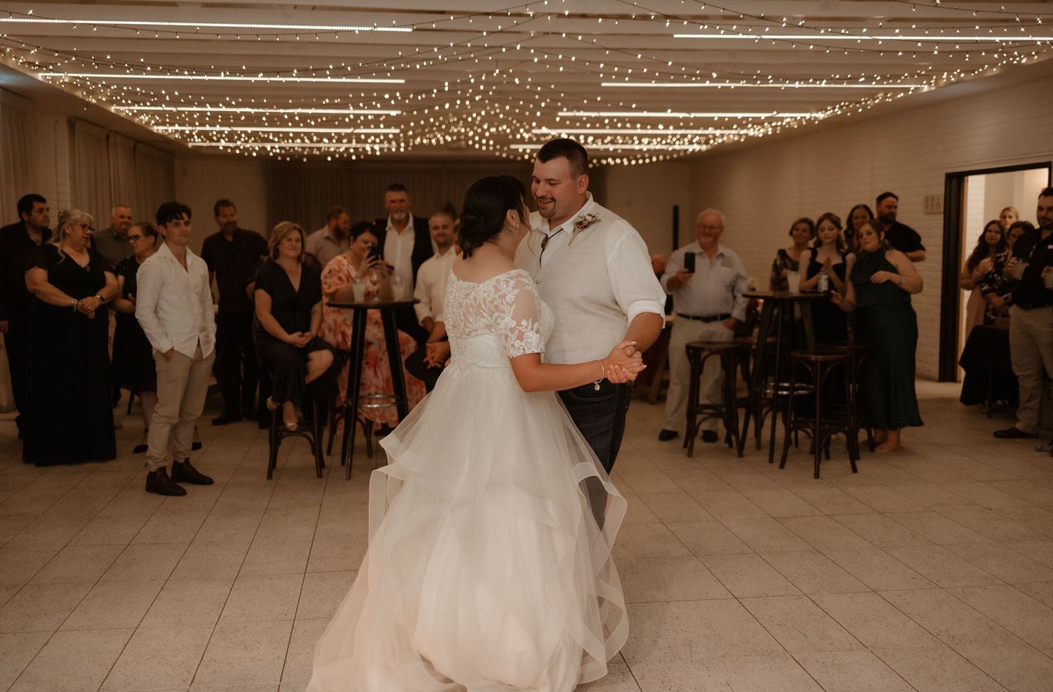 Bride in white tulle gown and groom share first dance under twinkling fairy lights at wedding reception.