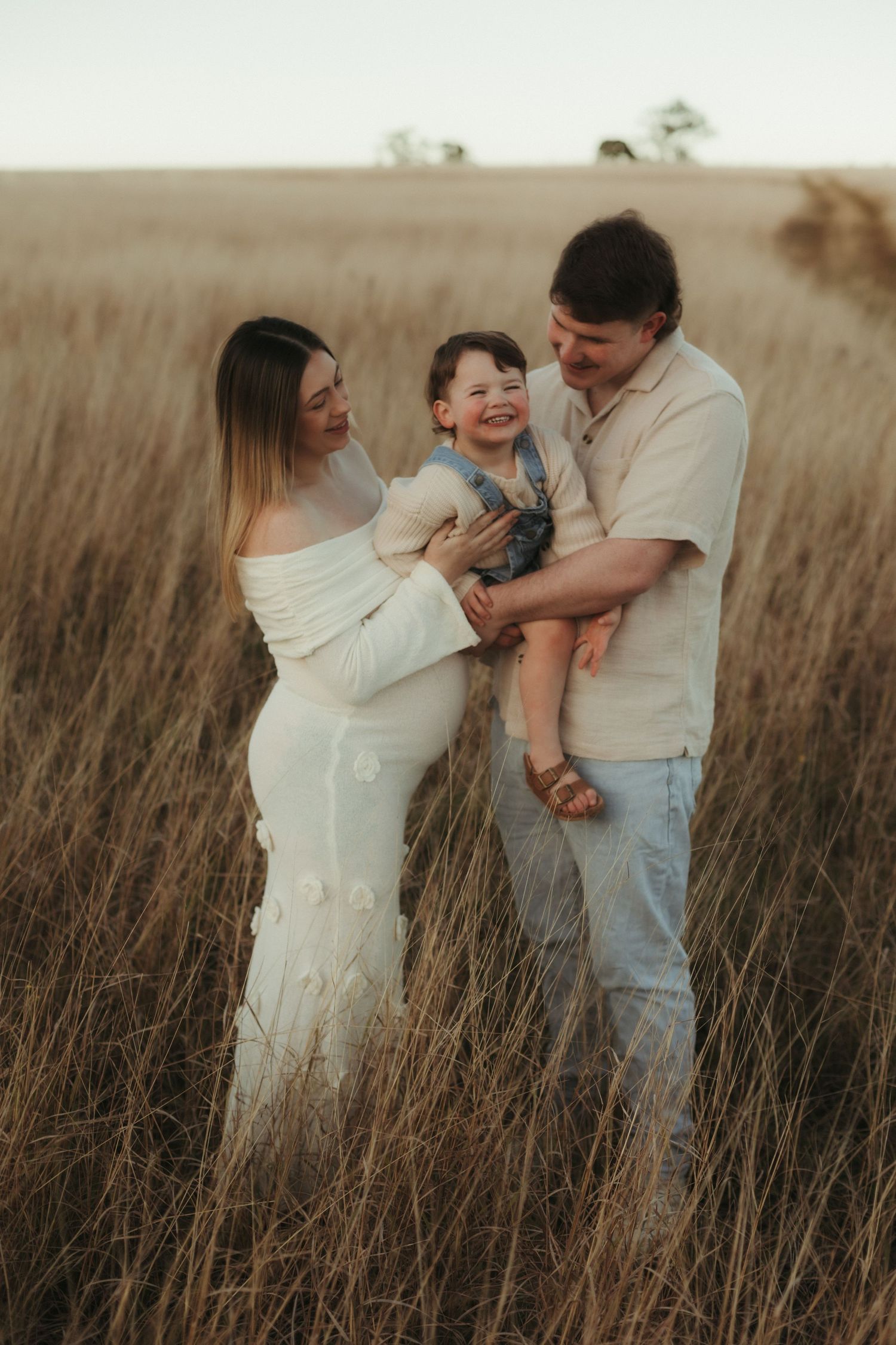Couple holding laughing toddler between them while standing in a golden grass field at sunset.