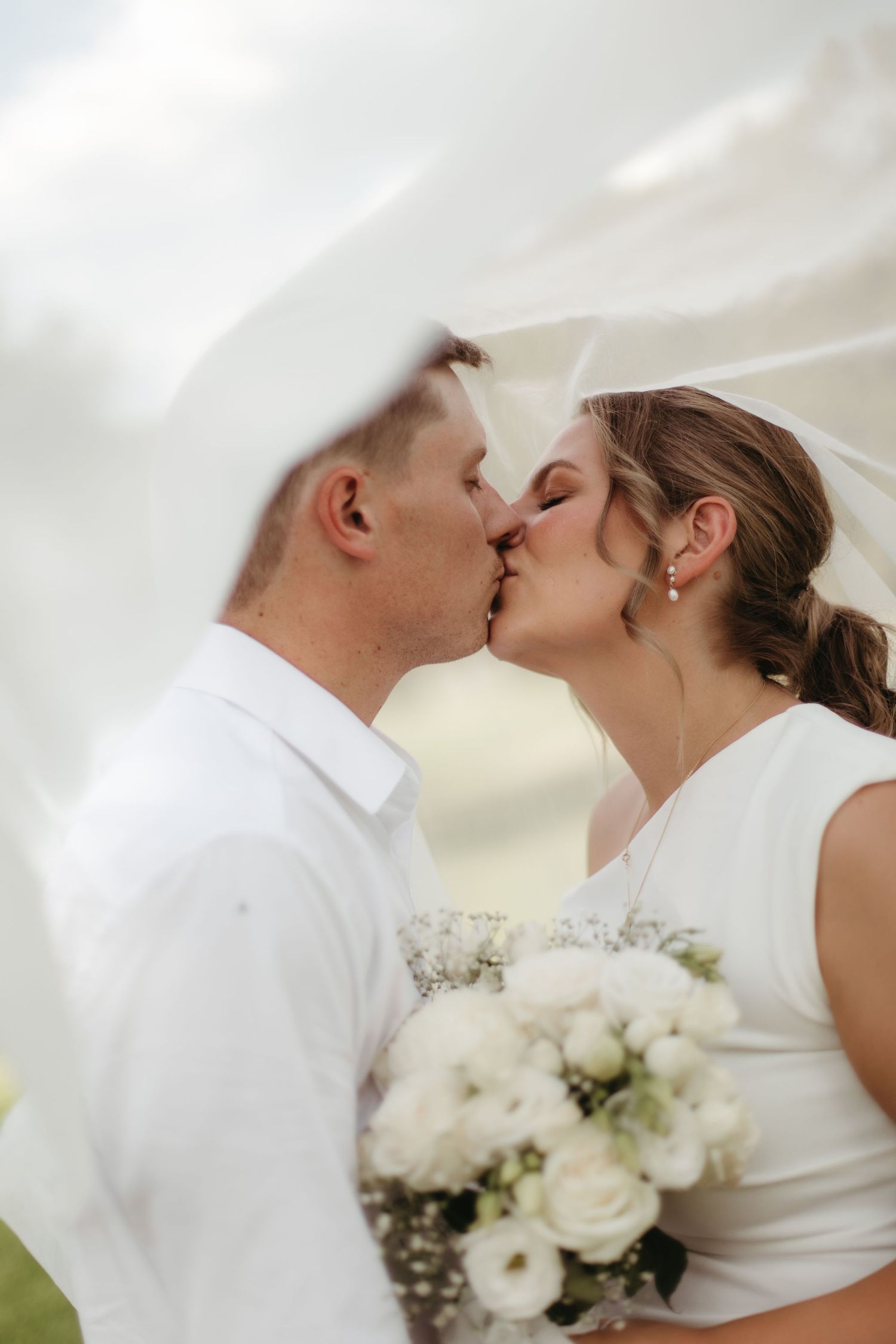 Bride and groom sharing first kiss under flowing white veil with white bouquet.