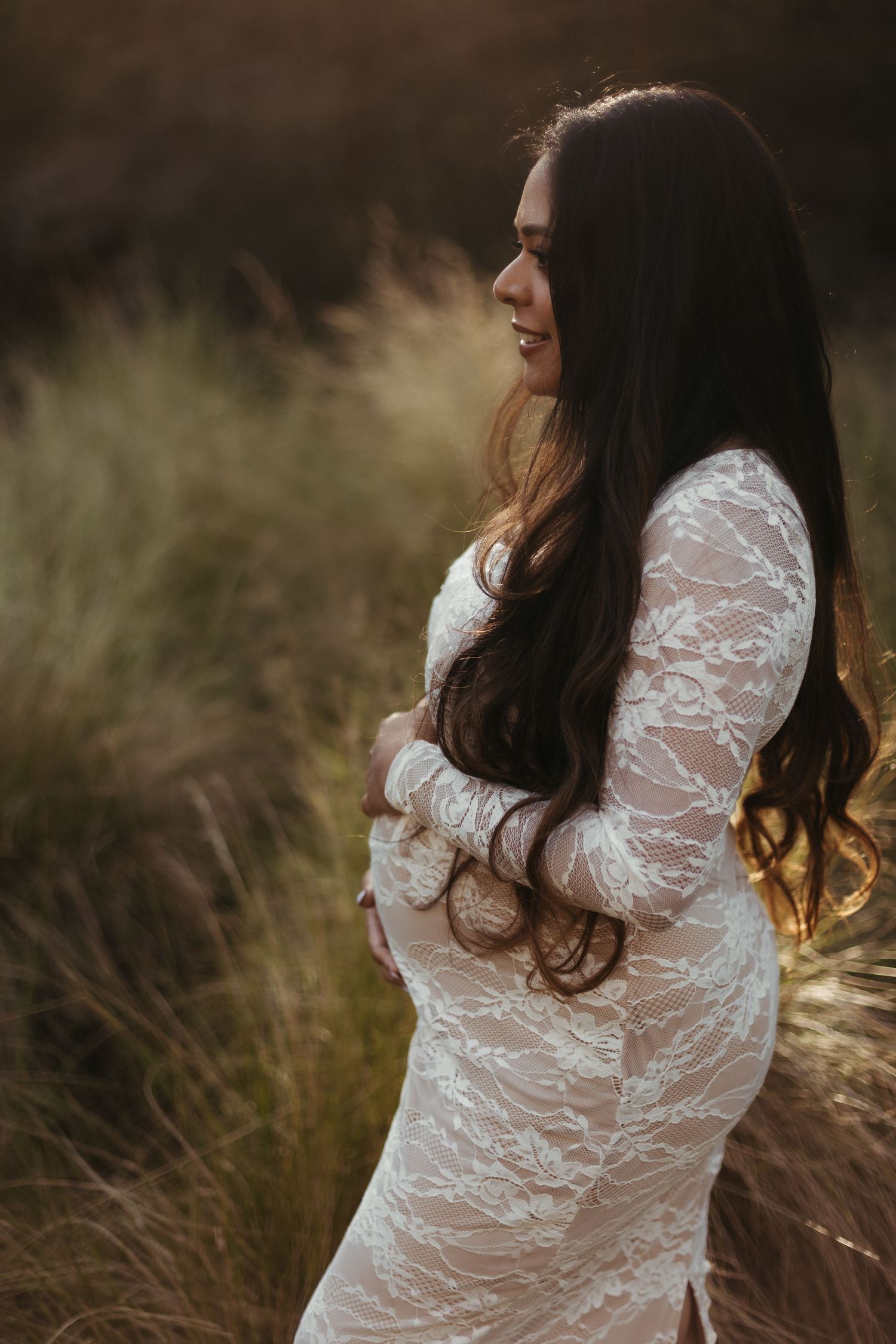 Pregnant woman in white lace dress holding baby bump in golden grass field at sunset.
