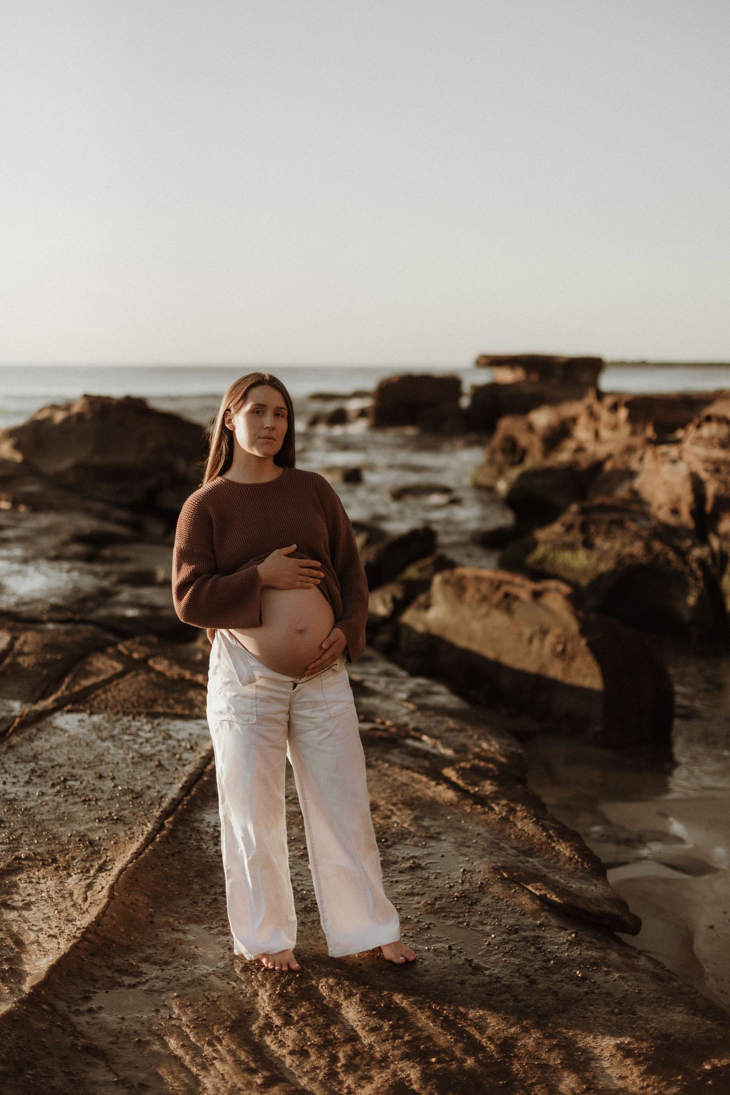 Pregnant woman in brown sweater and white pants standing on coastal rocks at sunset.