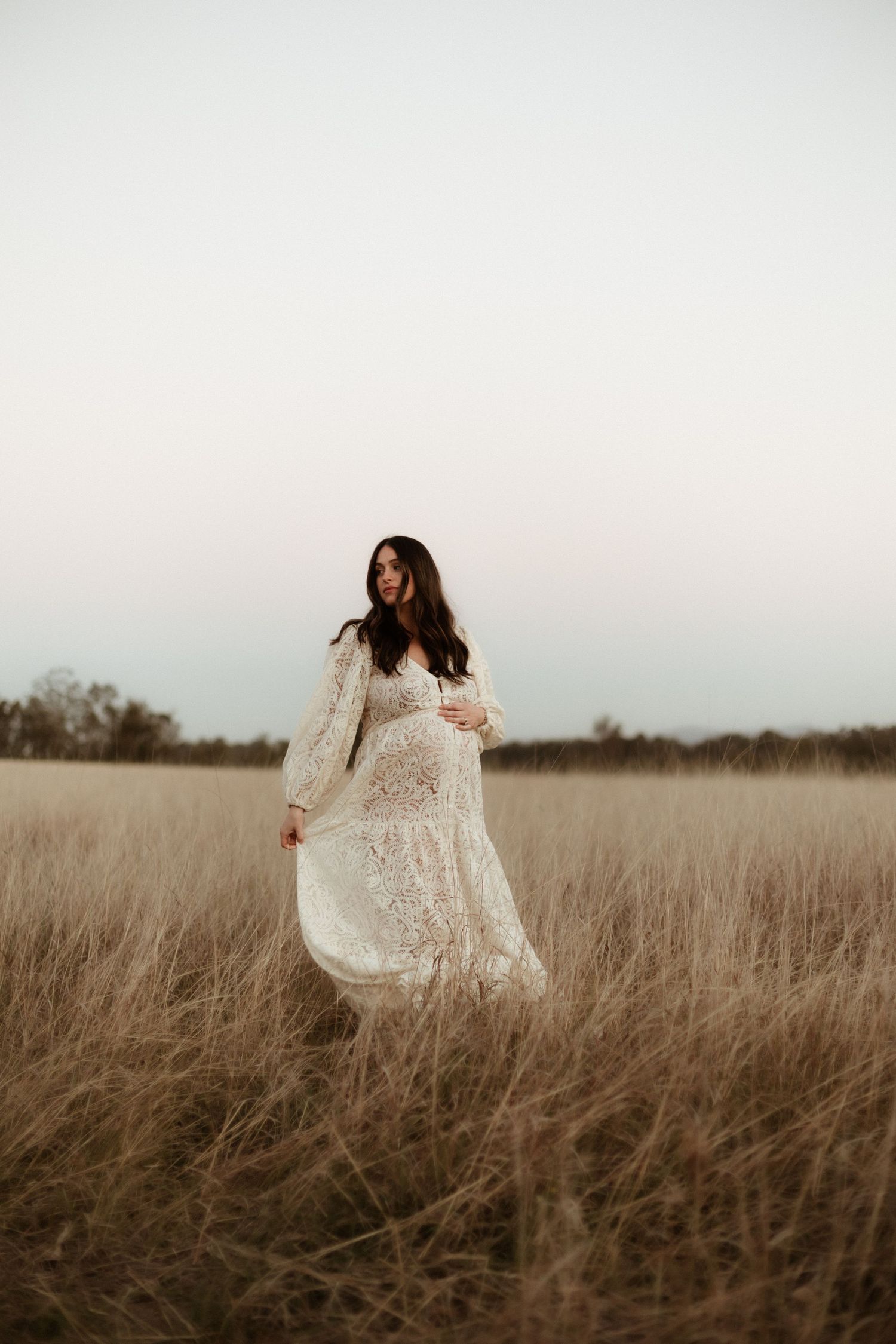 Pregnant woman in flowing white lace dress standing in golden wheat field at dusk.