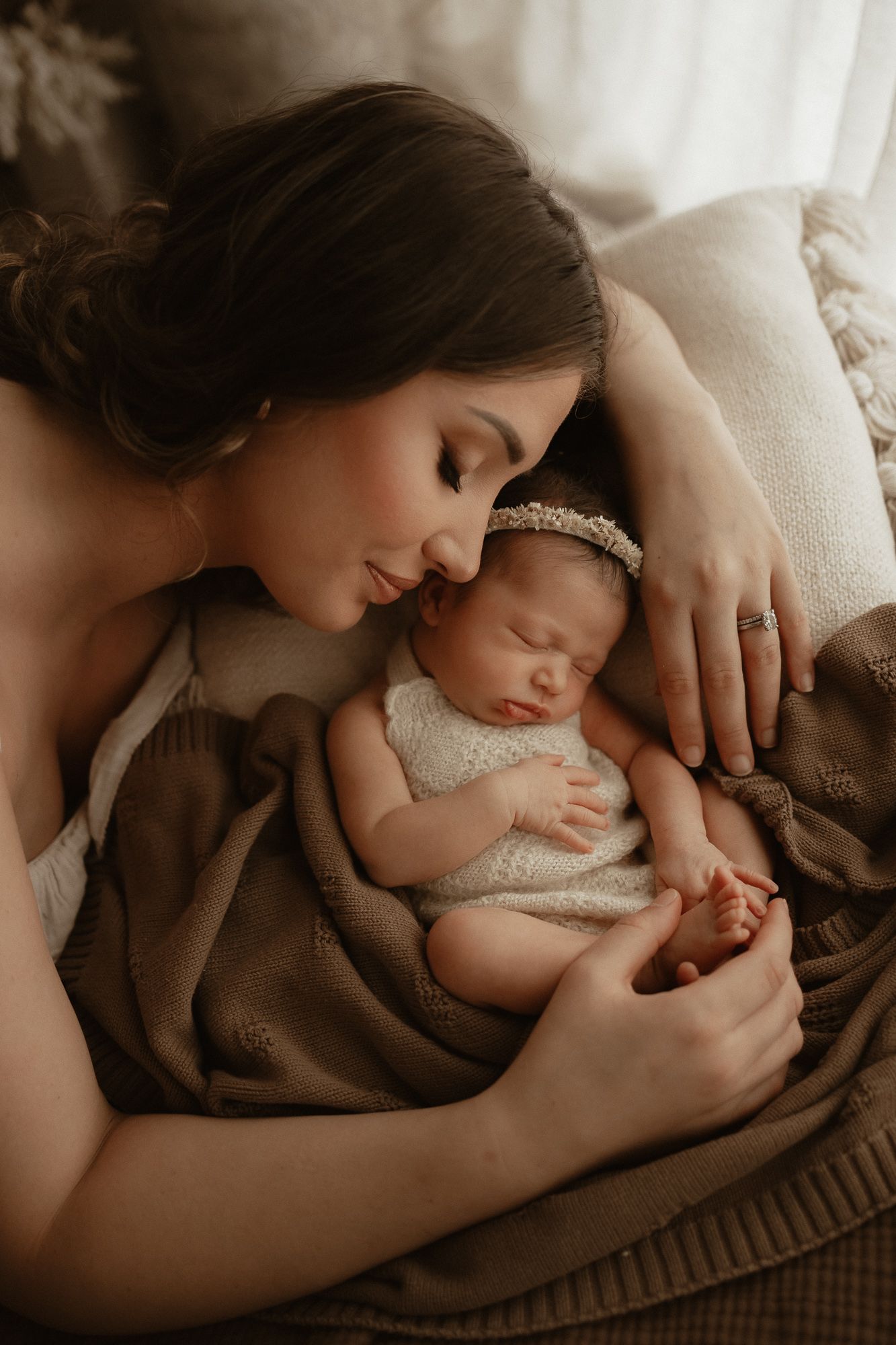 A mother tenderly nuzzles her sleeping newborn baby wrapped in a soft brown blanket with a delicate headband.