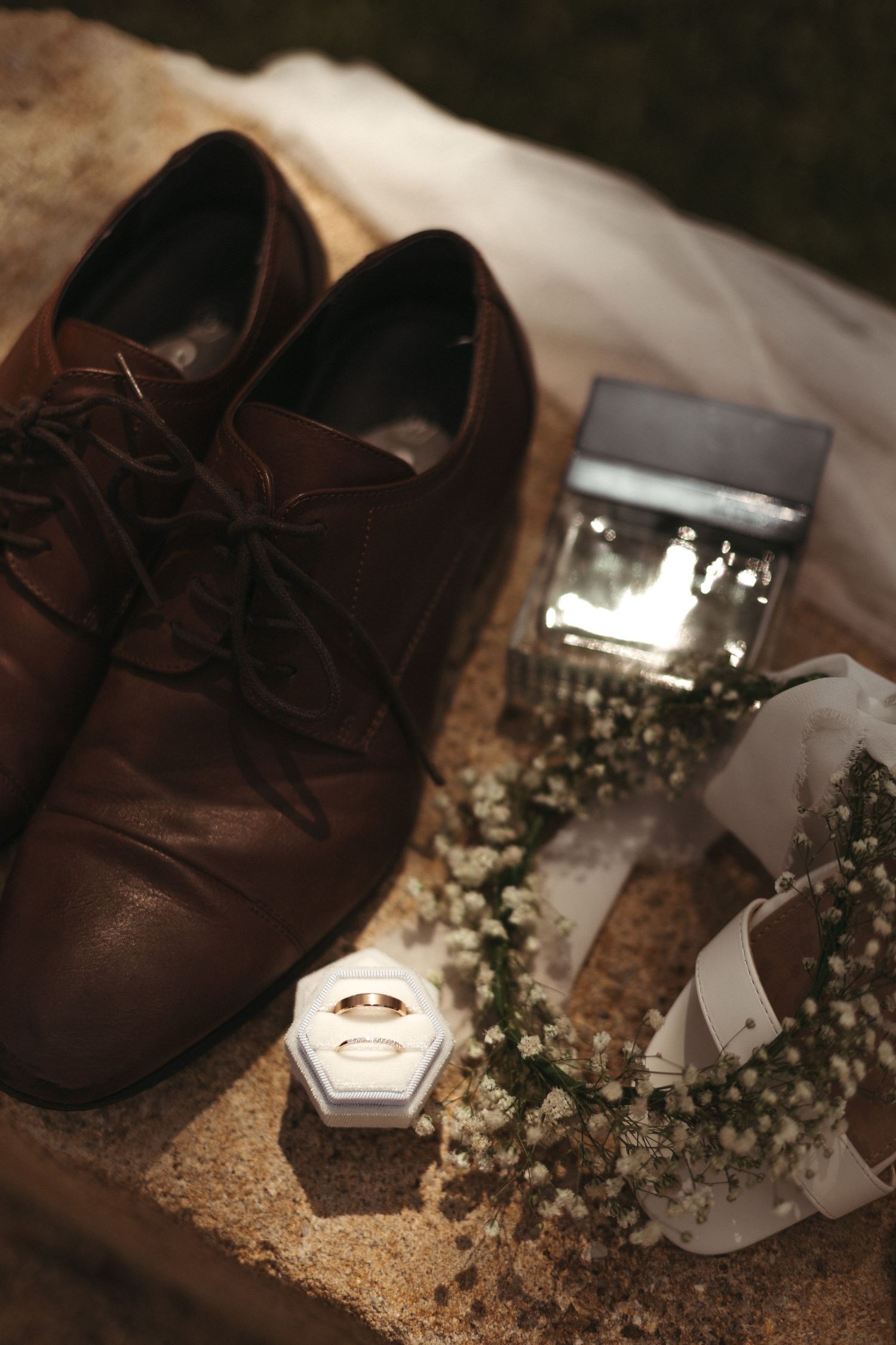 Brown leather dress shoes beside wedding rings in a box, baby's breath floral wreath, and cologne on a stone surface.