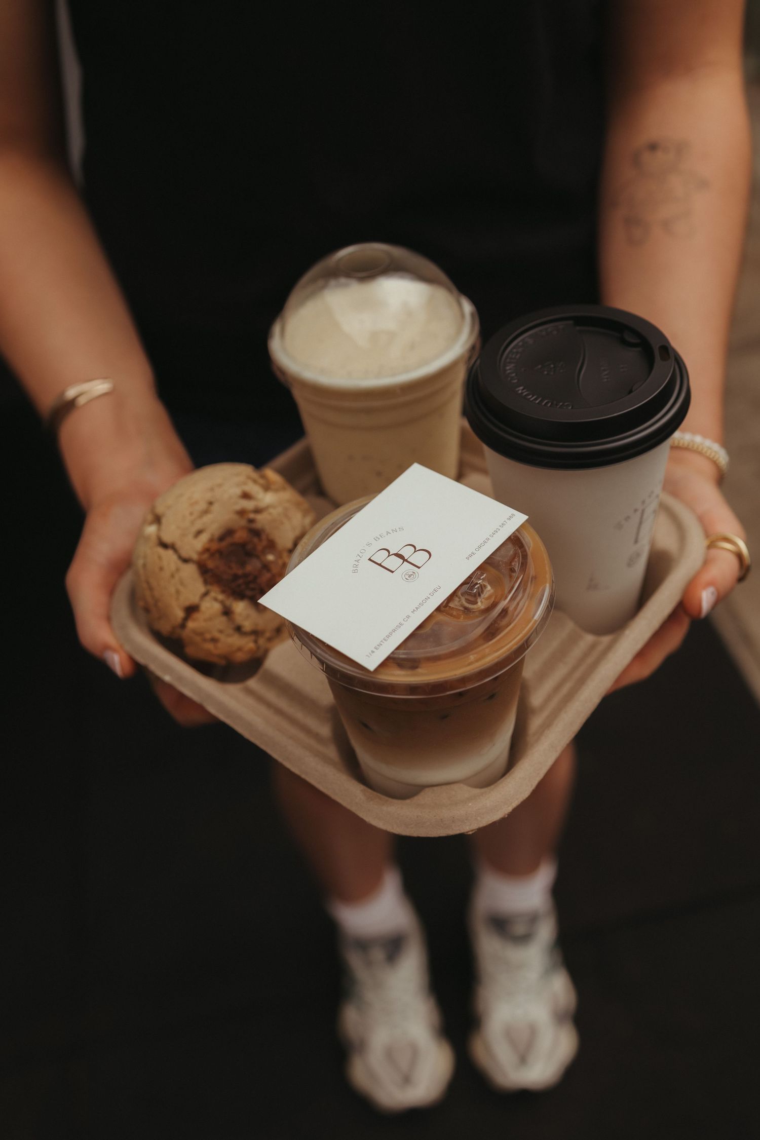 Hands with tattoos holding wooden tray of iced latte, hot coffees, cookies, and BB business card.