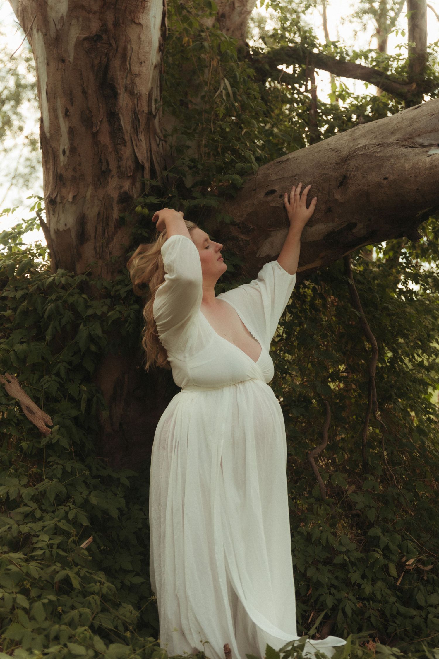 A pregnant woman in a white dress reaches up to touch a fallen tree trunk in a green forest.