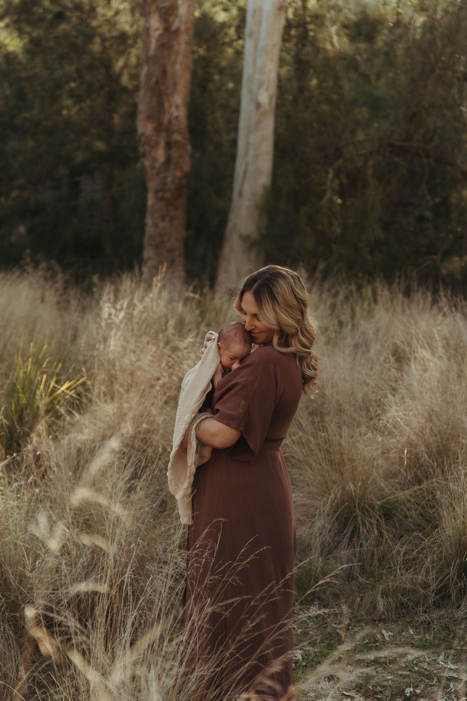 A mother in a brown dress holds her newborn baby wrapped in a cream blanket in a golden grass field.