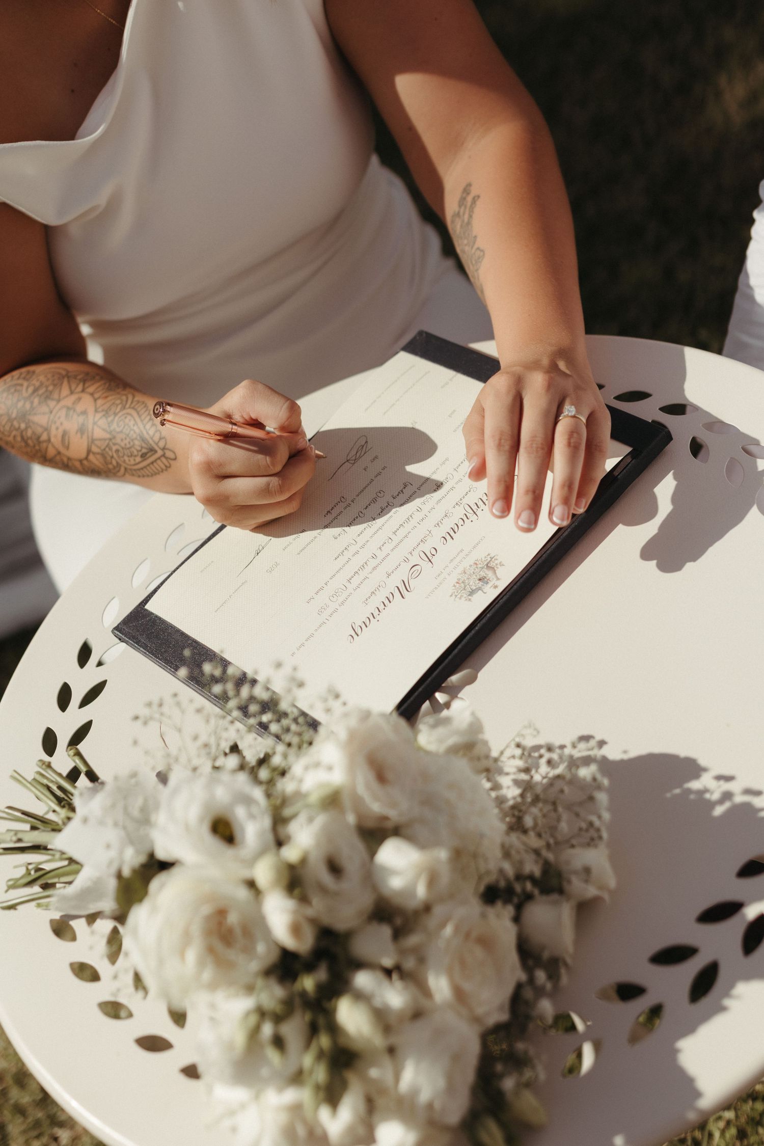 Bride signing marriage certificate at white table with white floral bouquet in foreground.