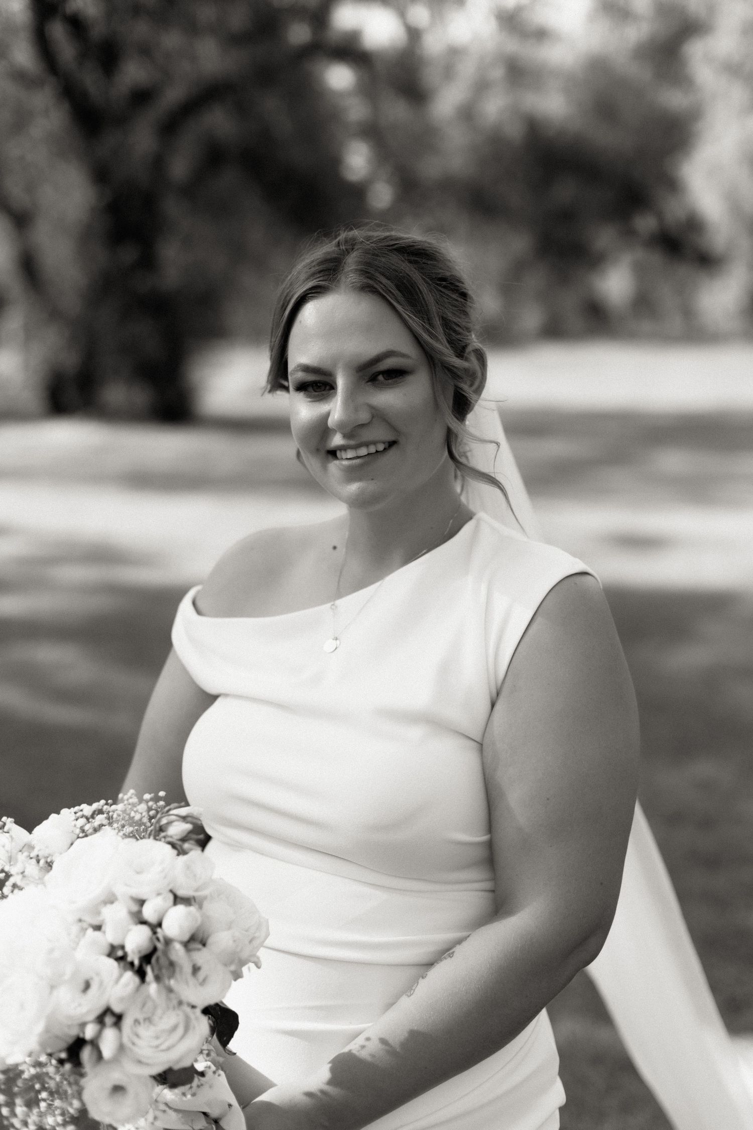 Smiling bride in white off-shoulder gown holding floral bouquet, elegant black and white outdoor portrait.