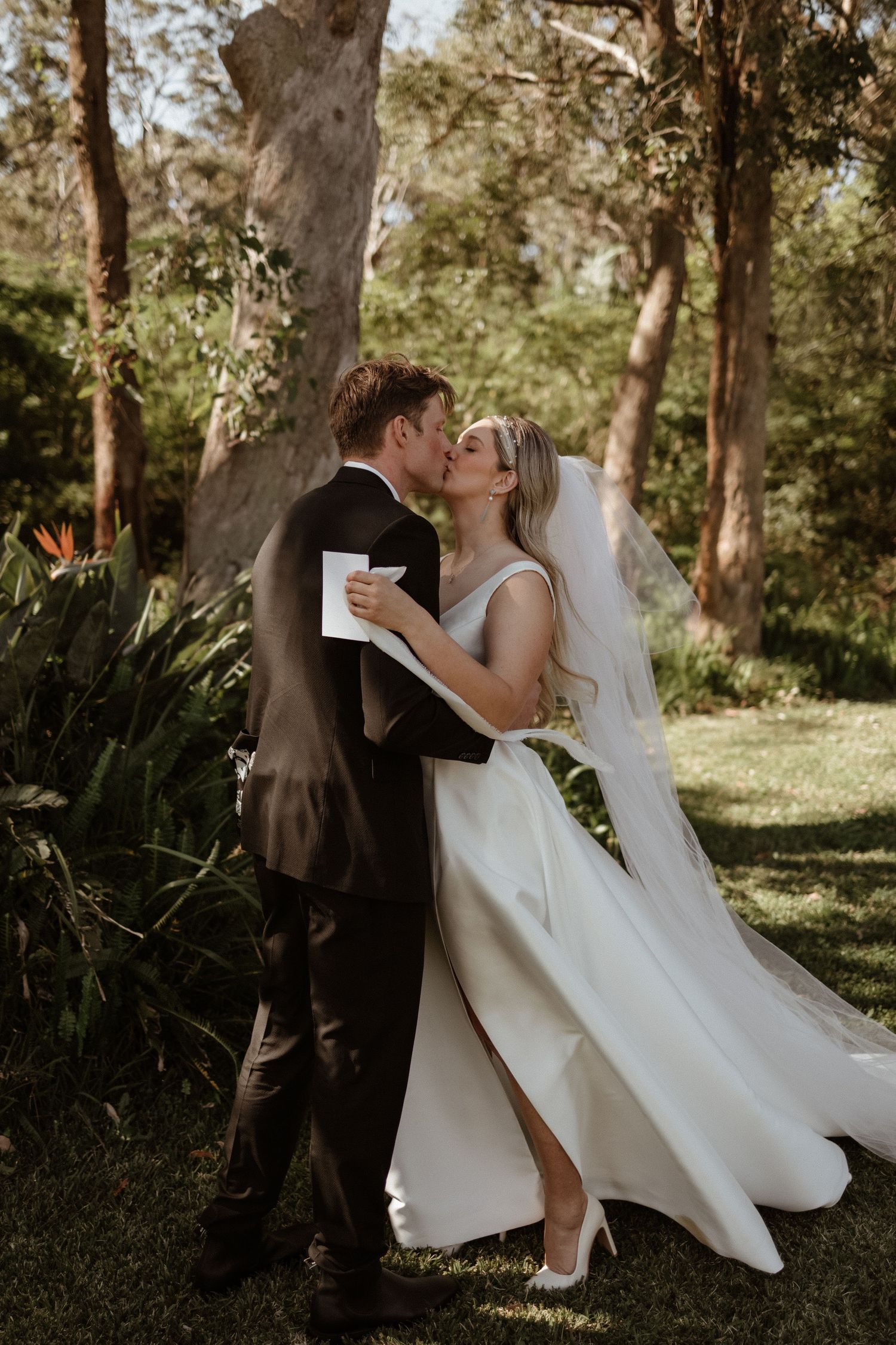Bride and groom share a romantic first kiss as newlyweds in a lush outdoor garden setting.