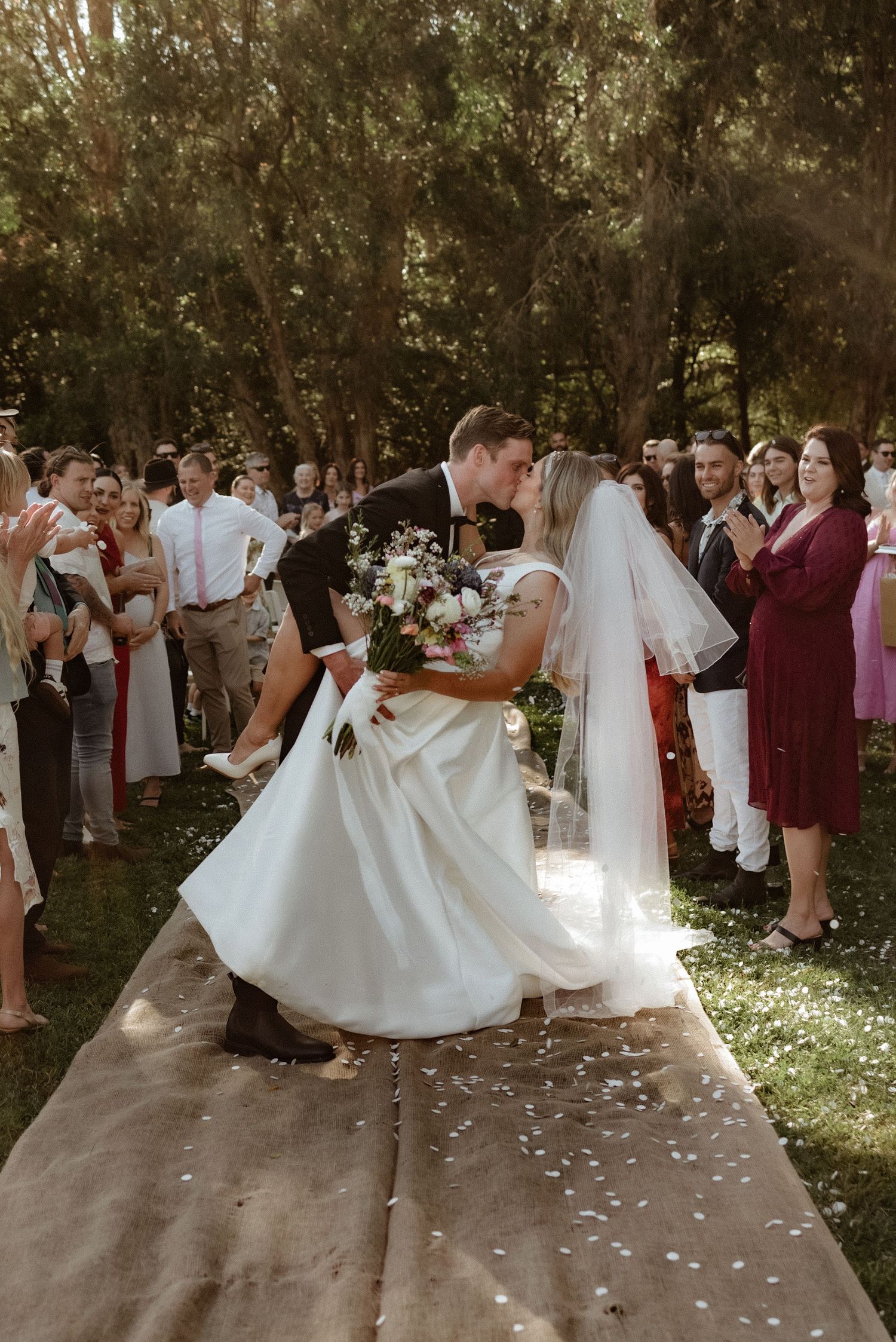 Groom dips bride for a romantic kiss on the aisle surrounded by cheering wedding guests.