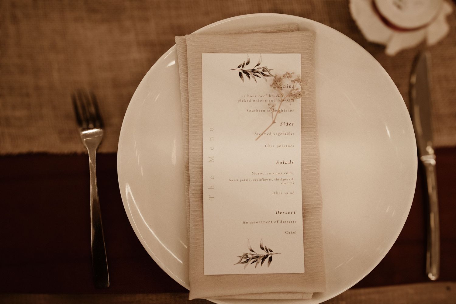 Elegant wedding menu card with botanical design placed on white plate with silverware at reception table.