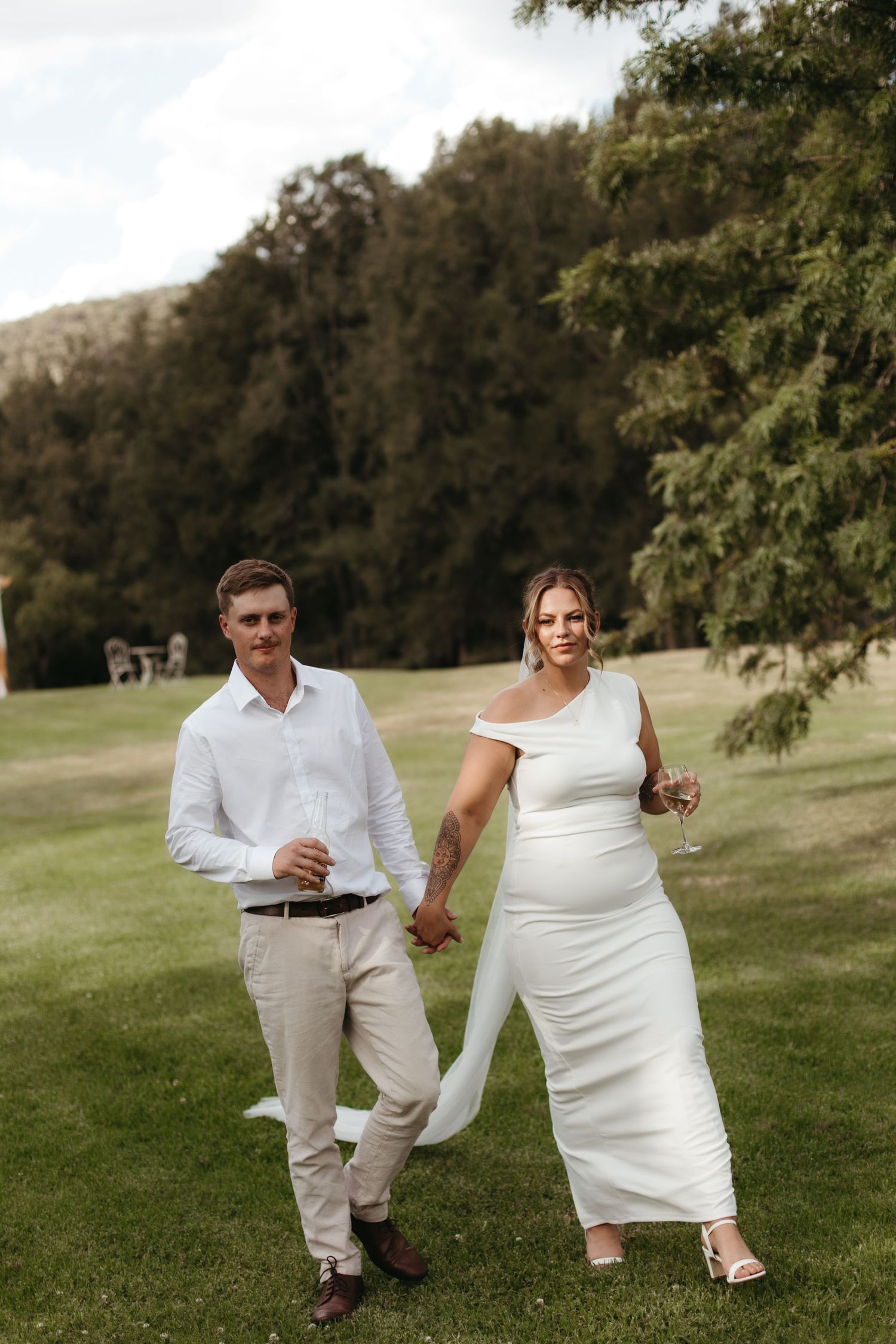 Bride and groom walking hand in hand on green lawn holding champagne glasses.