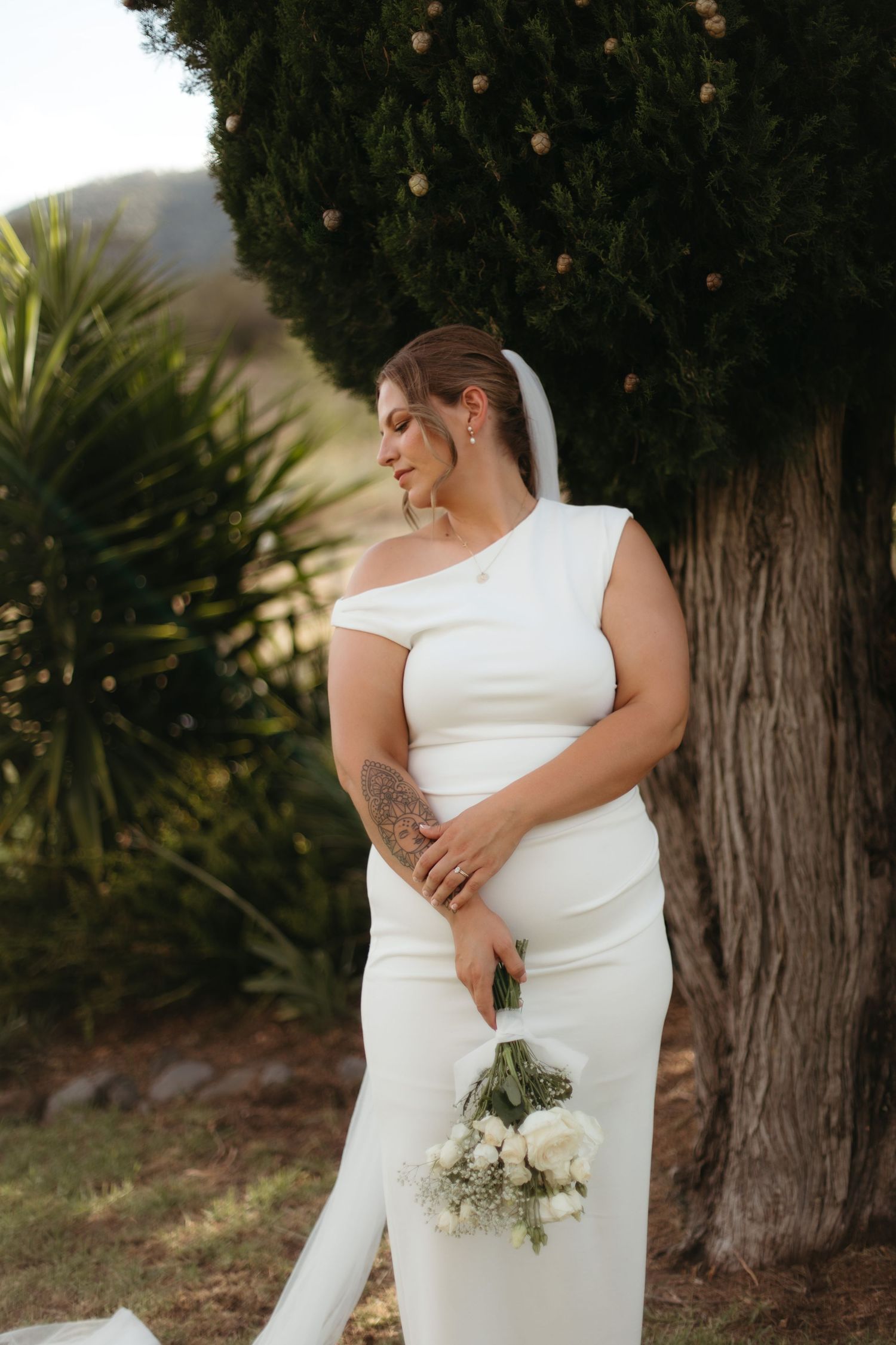 Bride in white one-shoulder gown holding bouquet of white roses, standing by tree with veil and arm tattoo.