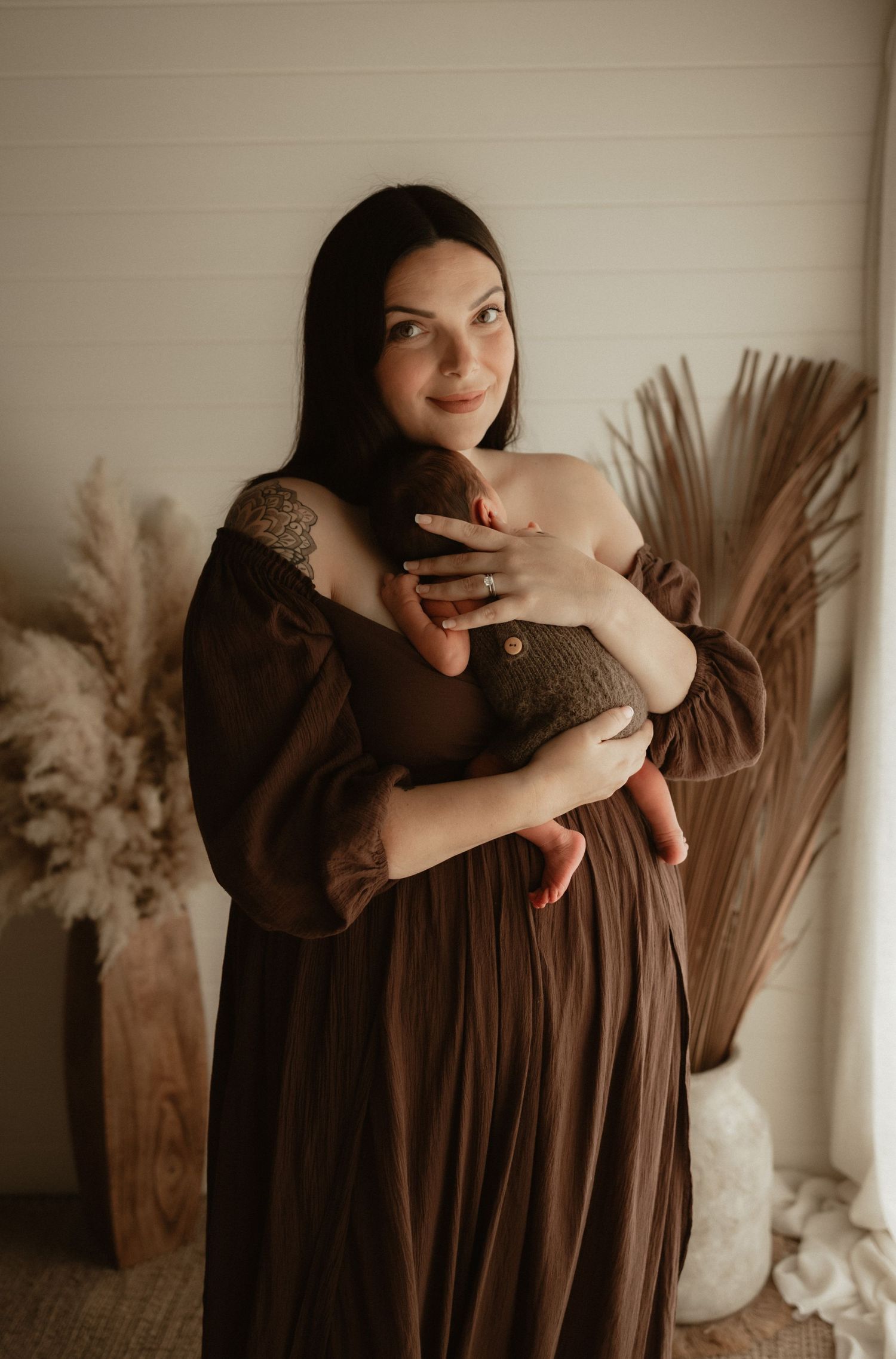 A brunette mother in a brown dress smiles while cradling her newborn baby against her chest.