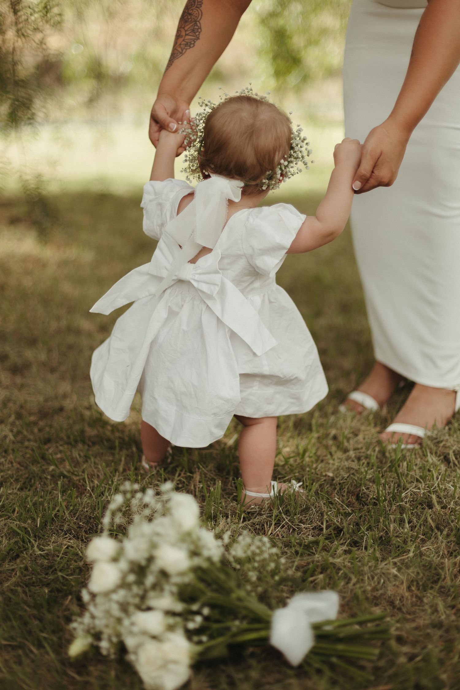 Adorable baby girl in white dress with bow and floral crown walking with bride at outdoor wedding.