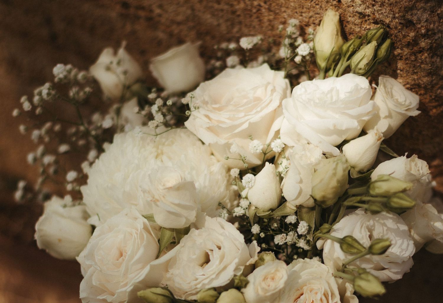 Elegant white wedding bouquet featuring roses, lisianthus, and baby's breath arranged on a rustic stone surface.