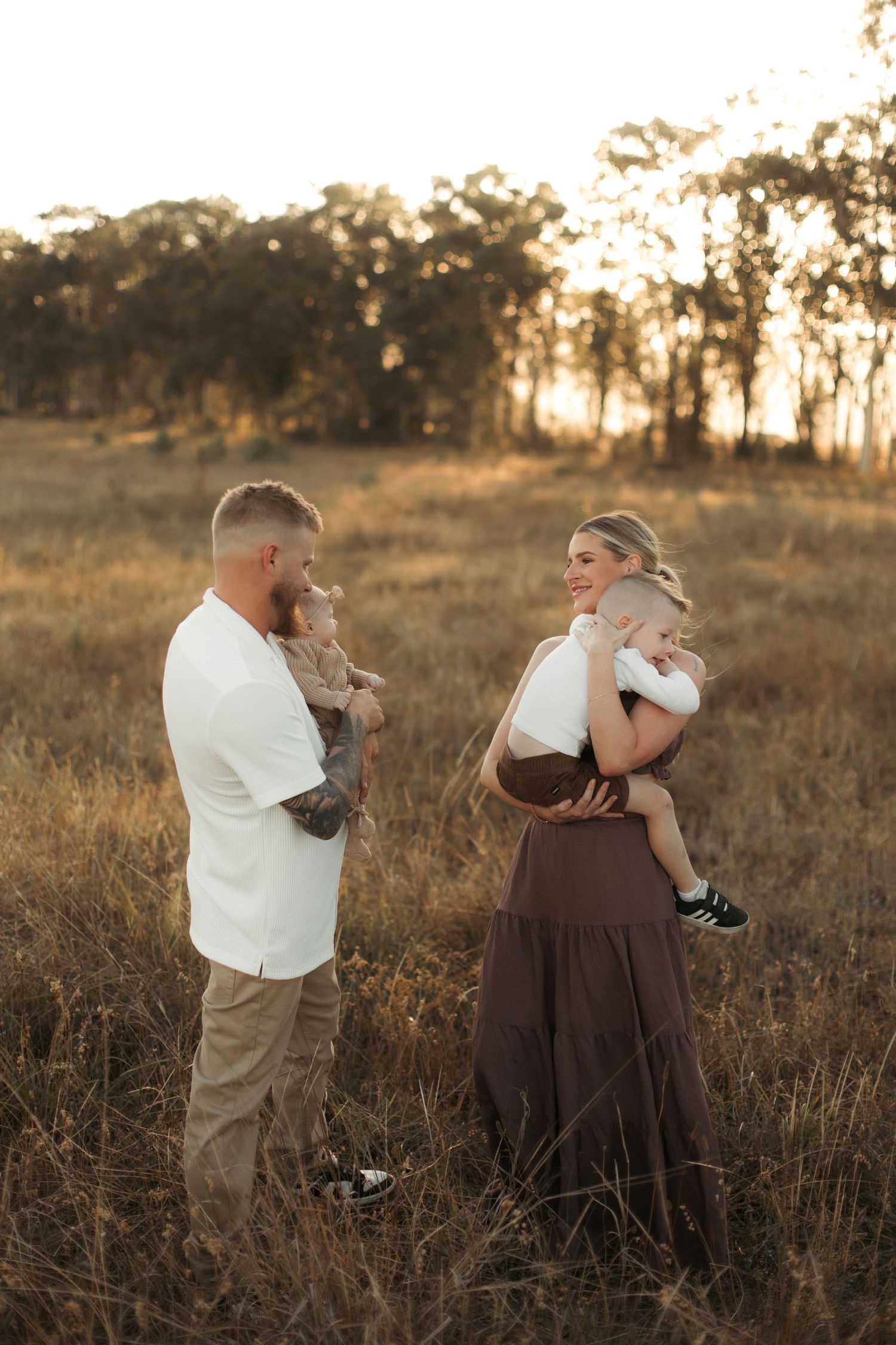Family of four sharing a warm golden hour moment in a sunlit field, parents holding young children outdoors.