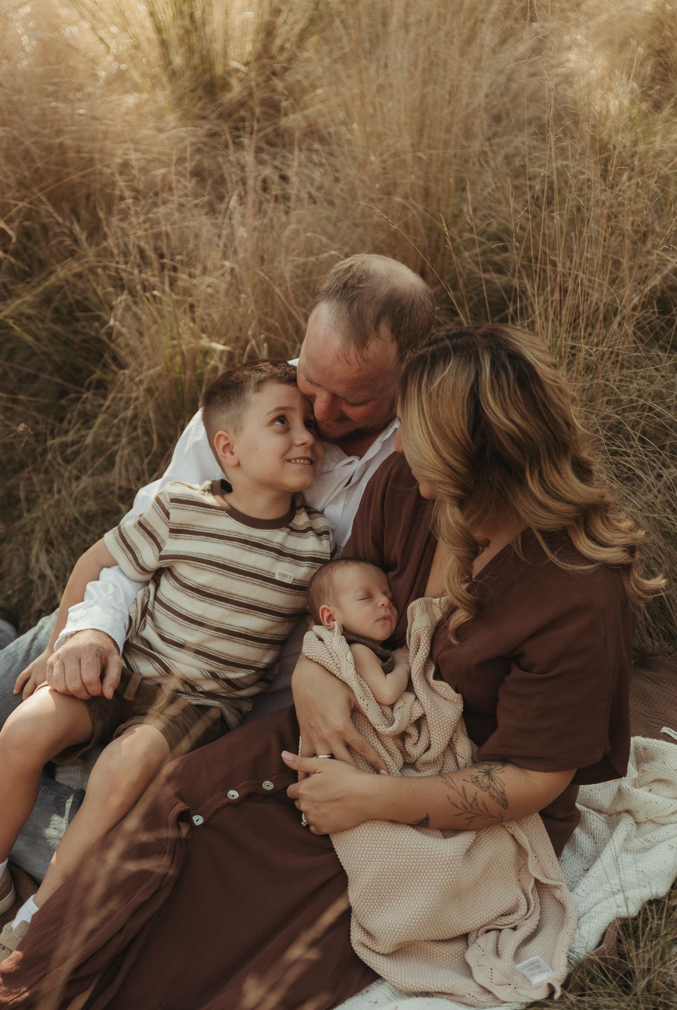 Family of four sits in golden grass field, parents gazing lovingly at newborn baby while young boy smiles warmly.