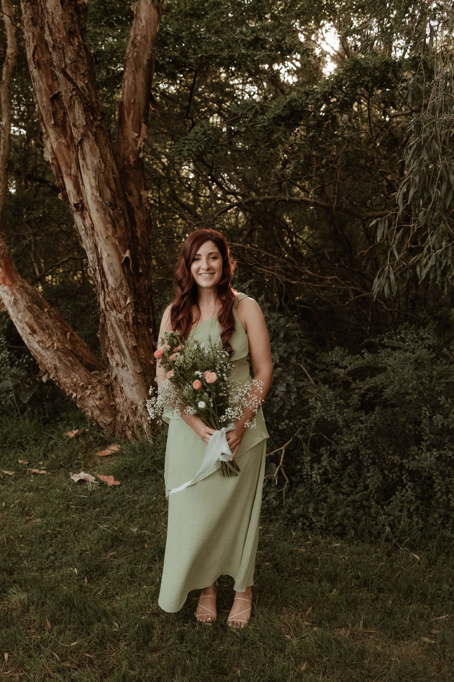 Smiling bridesmaid in sage green gown holding pink floral bouquet outdoors among lush green trees.