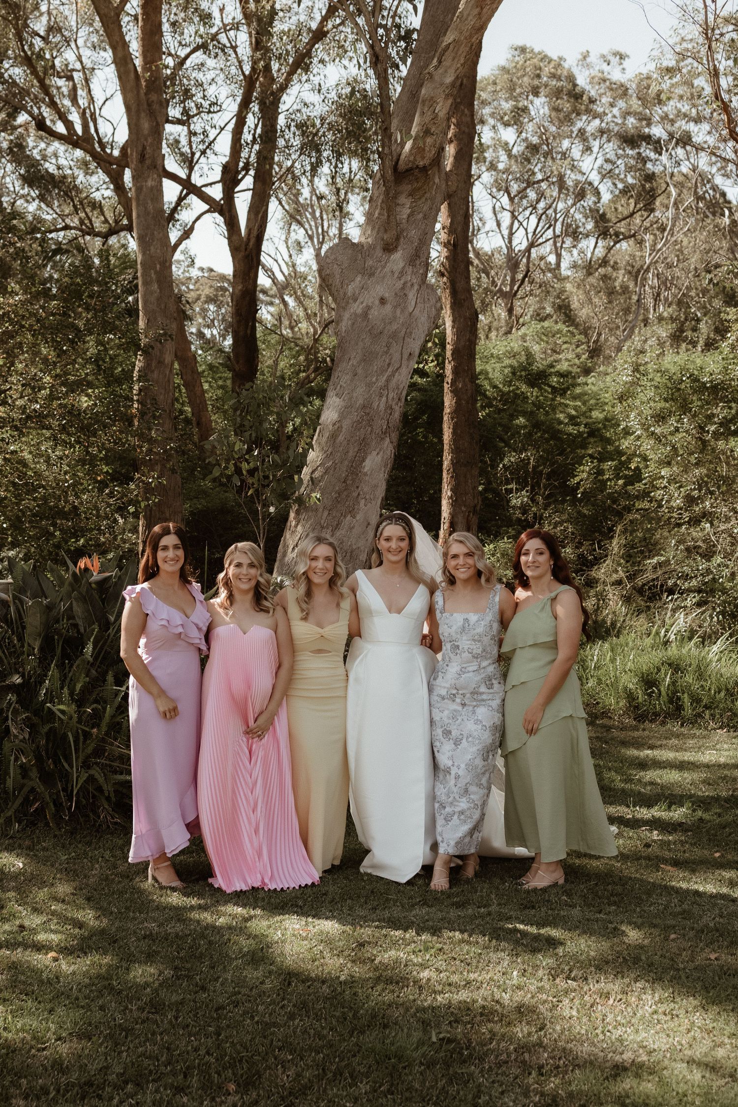 Bride poses with five bridesmaids in pastel gowns in a lush green outdoor garden setting.