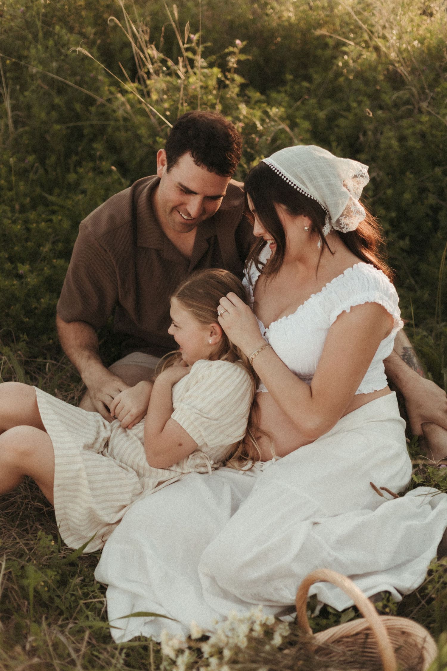 Happy family sitting in a meadow, parents smiling at their young daughter in warm golden light.