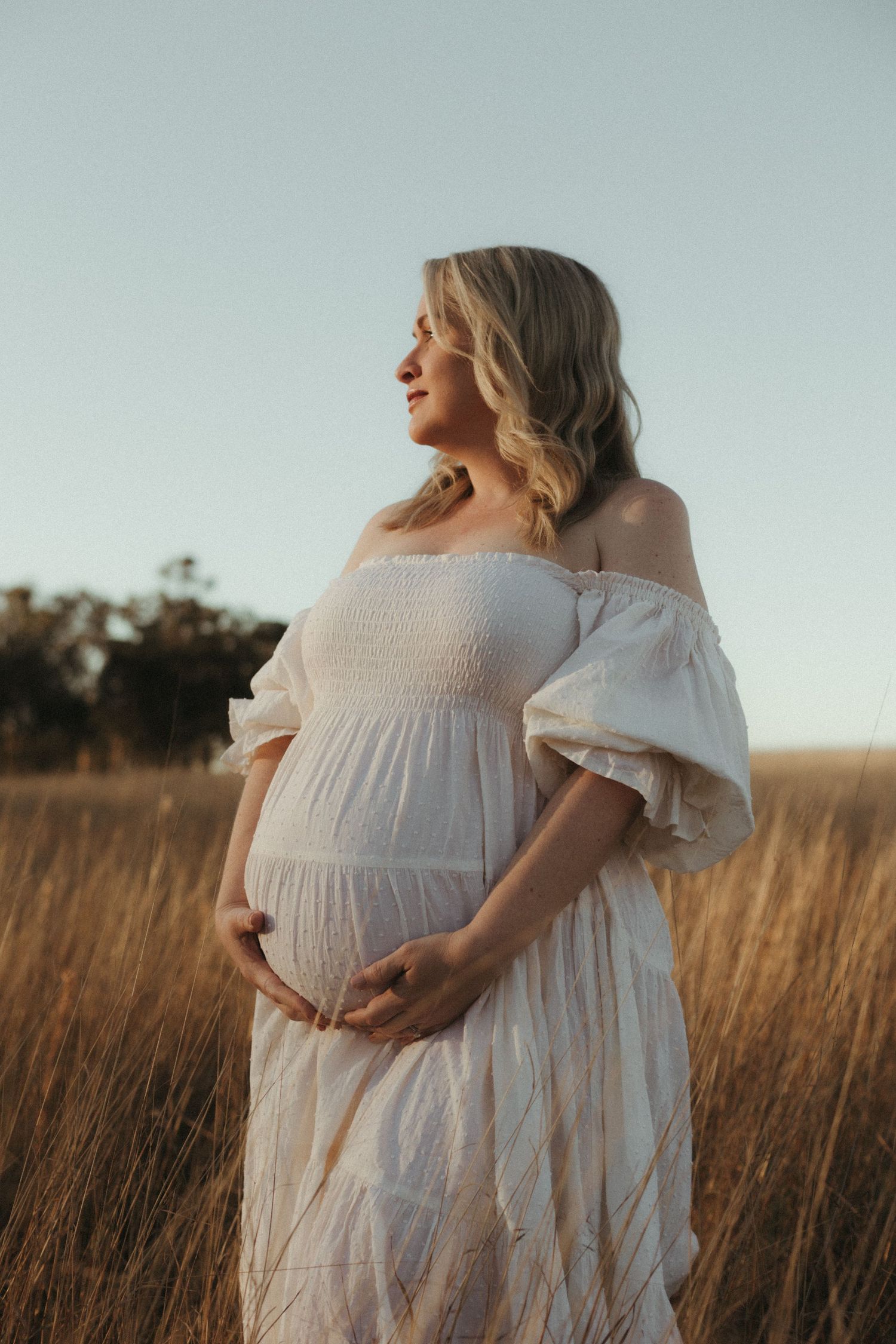 Blonde pregnant woman in white off-shoulder dress holding her baby bump in a golden field at dusk.