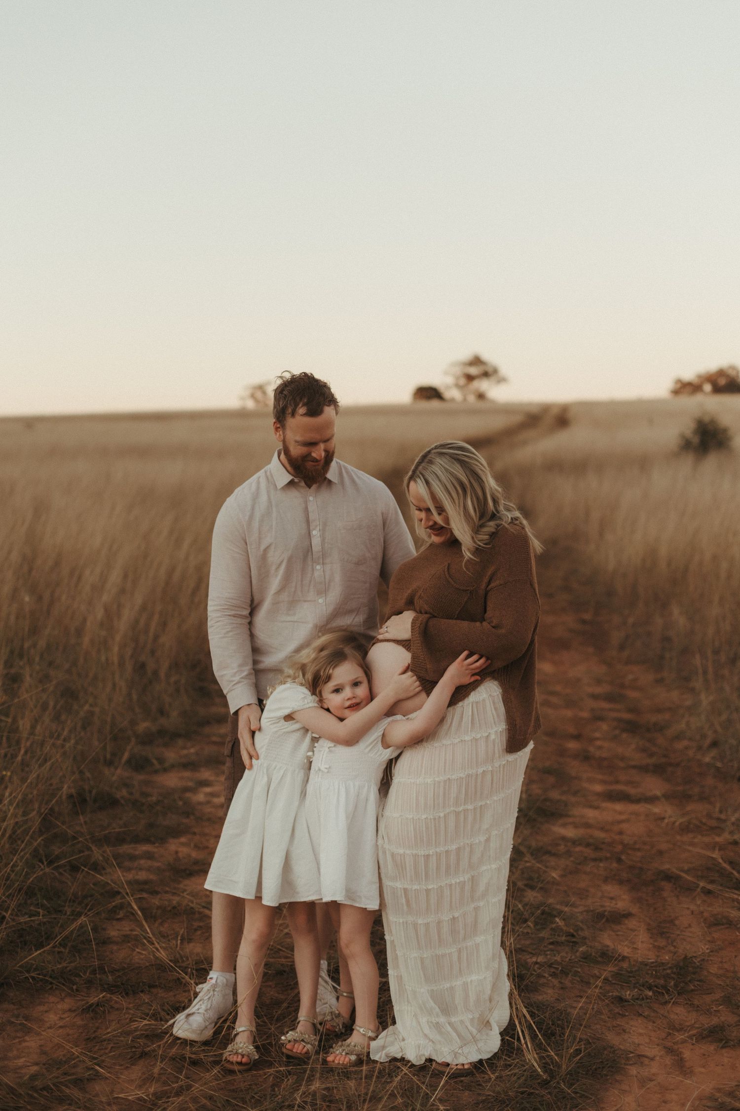 Family of three with bearded father, pregnant mother, and young daughter on a dirt path in a field.