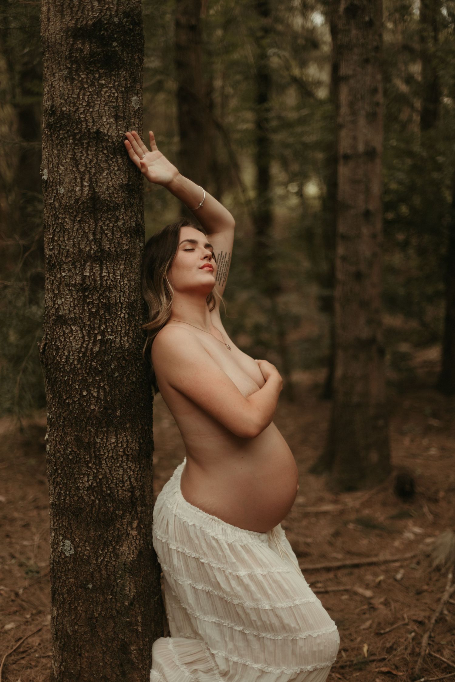 Topless pregnant woman in white ruffled skirt leans against forest tree, hand raised above head, eyes closed.