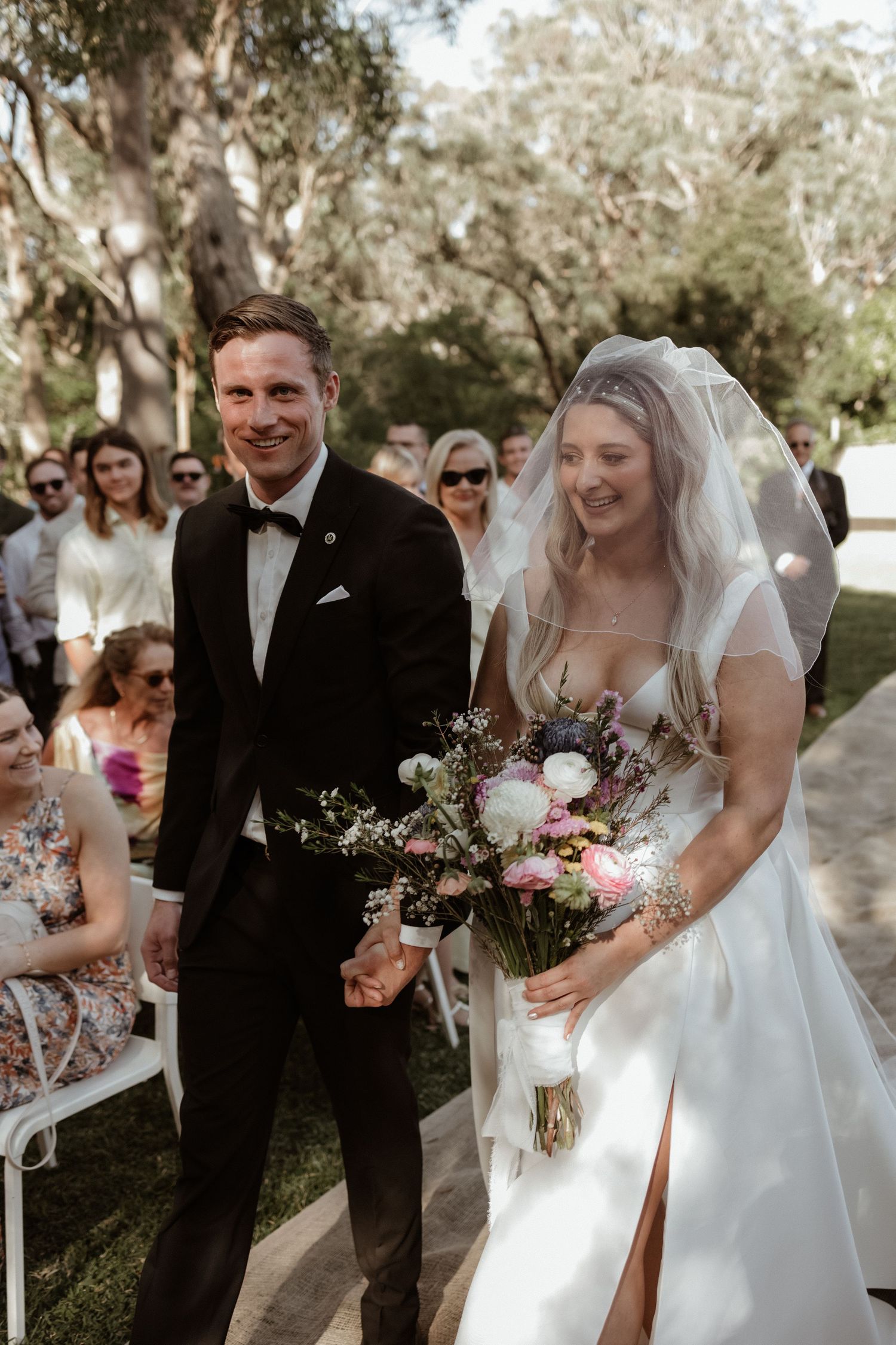 Smiling bride holding floral bouquet and groom in black tuxedo walking down outdoor wedding aisle.