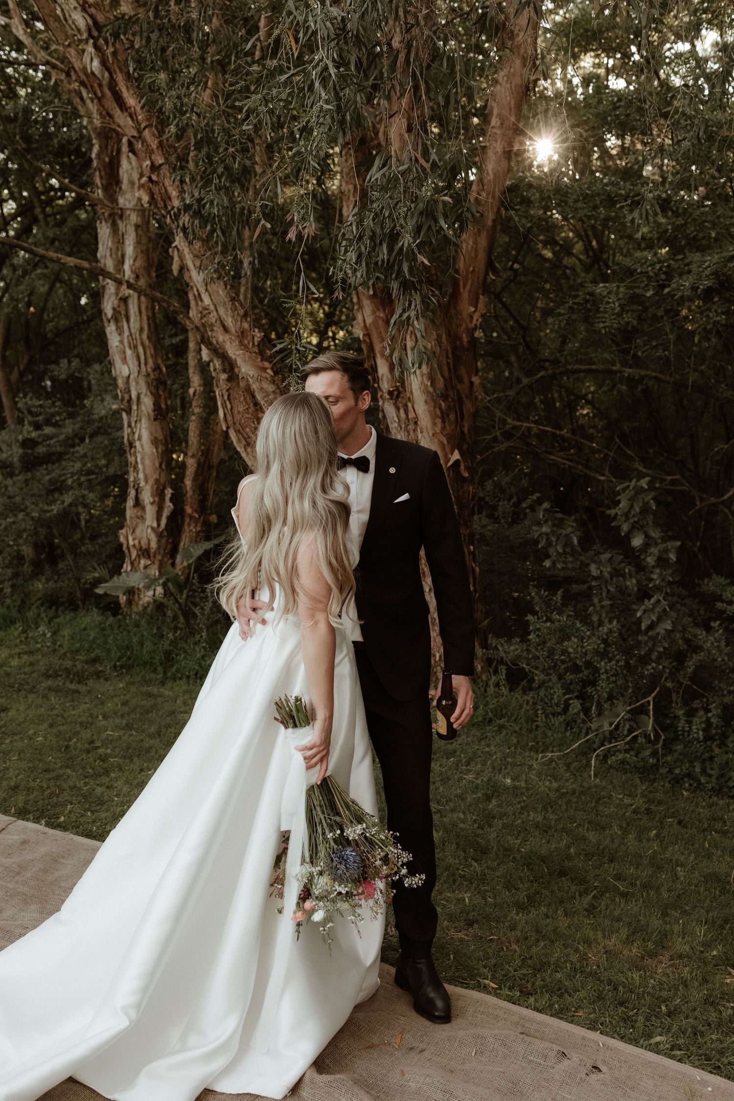 Romantic wedding couple embracing among tall eucalyptus trees, bride in white gown with long train.