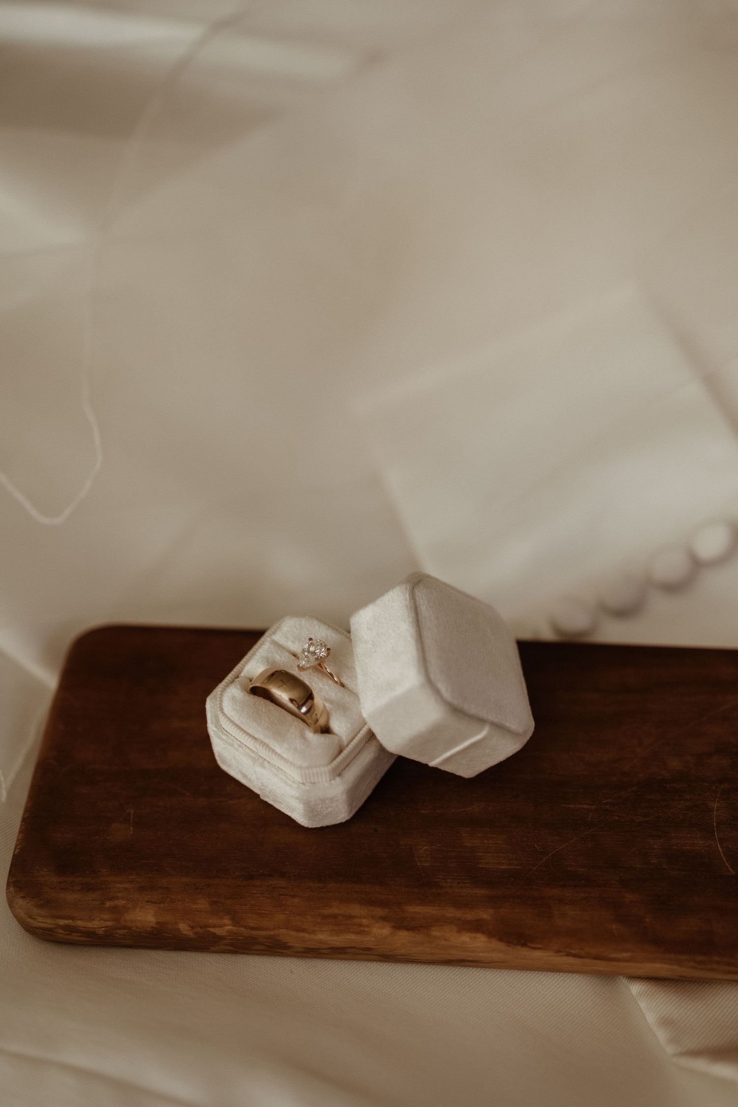 Gold wedding rings displayed in an open white velvet ring box on a rustic wooden board with soft white fabric.