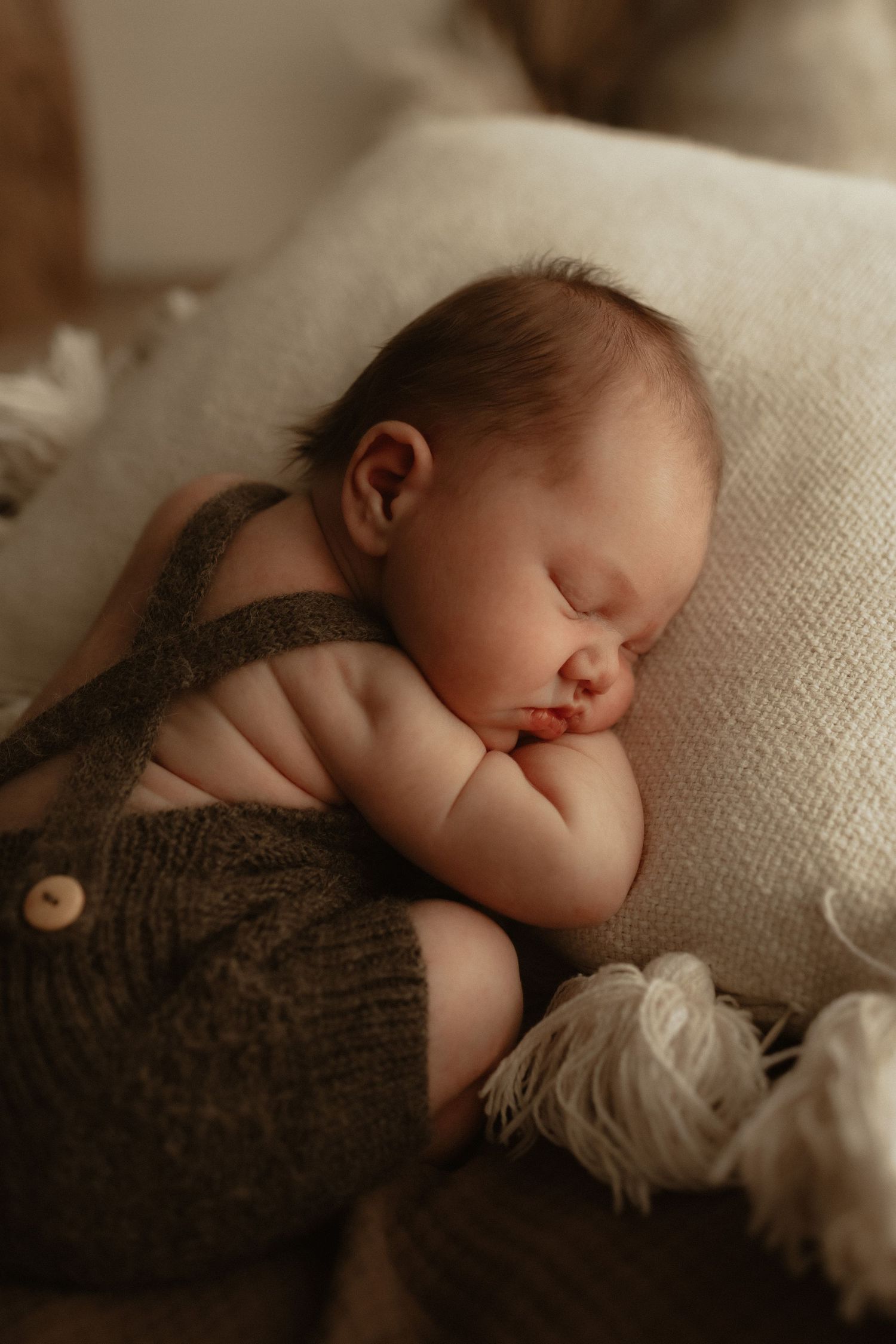 A peaceful newborn baby sleeping curled up on a soft blanket, wearing cozy brown knit overalls.
