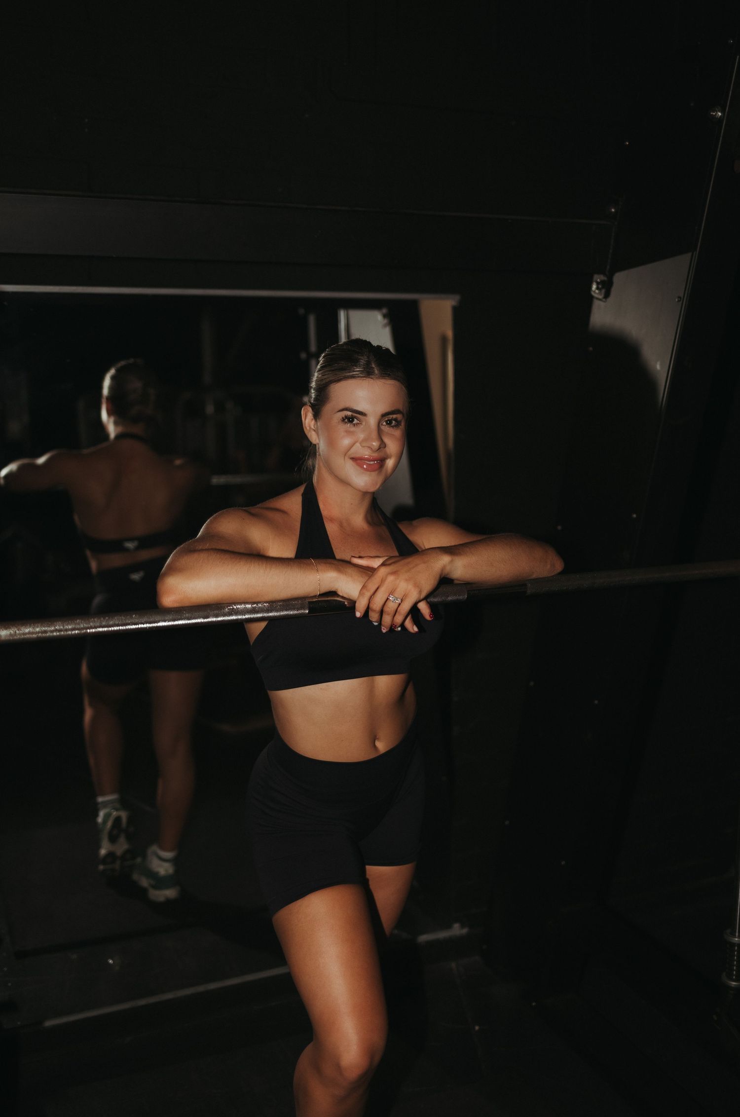 Fit woman in black sports bra and shorts leaning on gym barre smiling at camera.