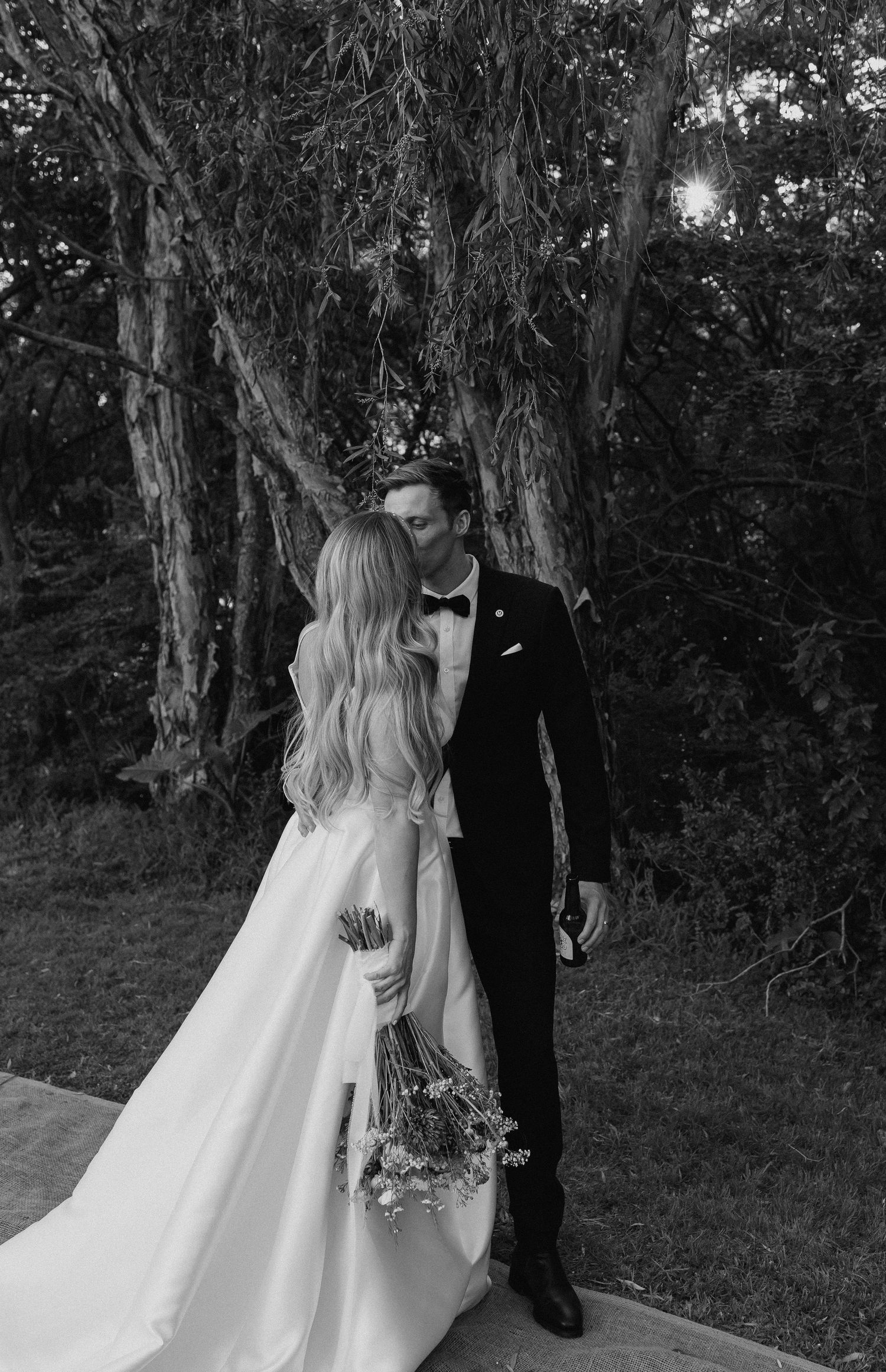 Black and white photo of a bride and groom sharing a kiss outdoors, surrounded by trees, bride holding a wildflower bouquet.