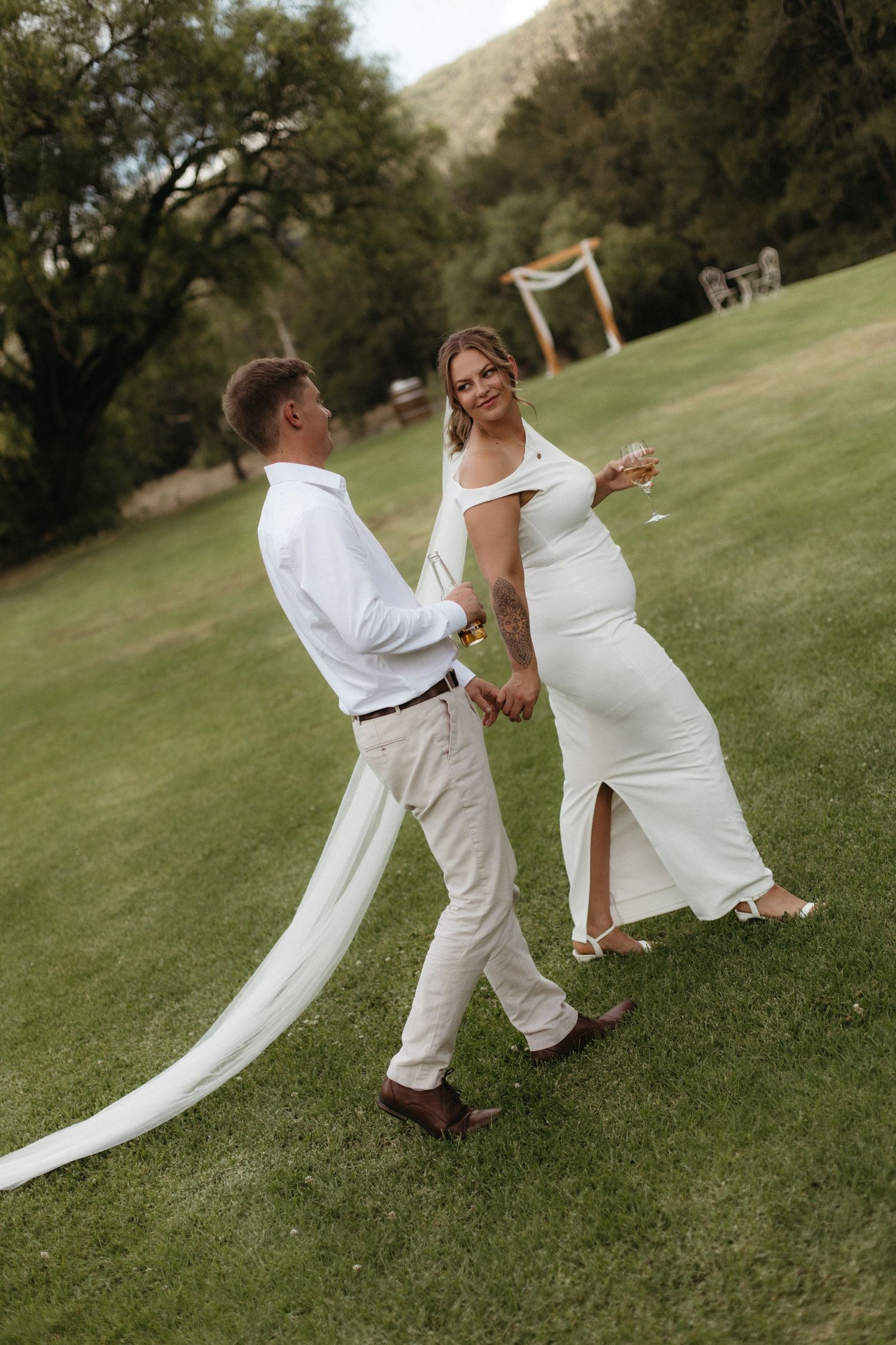 Bride and groom walking hand in hand on green lawn, bride in white fitted gown with veil, groom in white shirt.