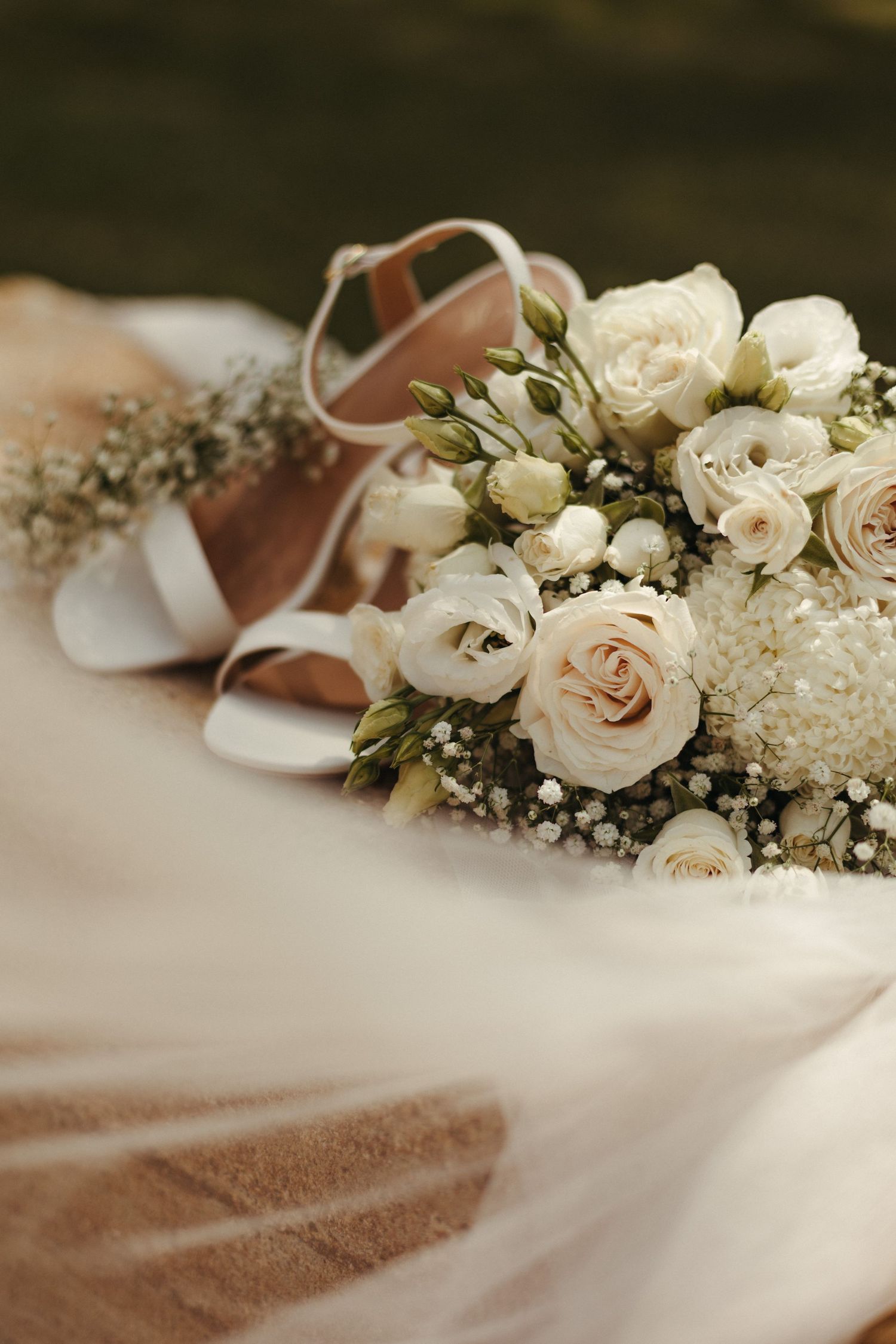 Bridal bouquet of white roses and gypsophila beside elegant wedding heels on a soft veil.