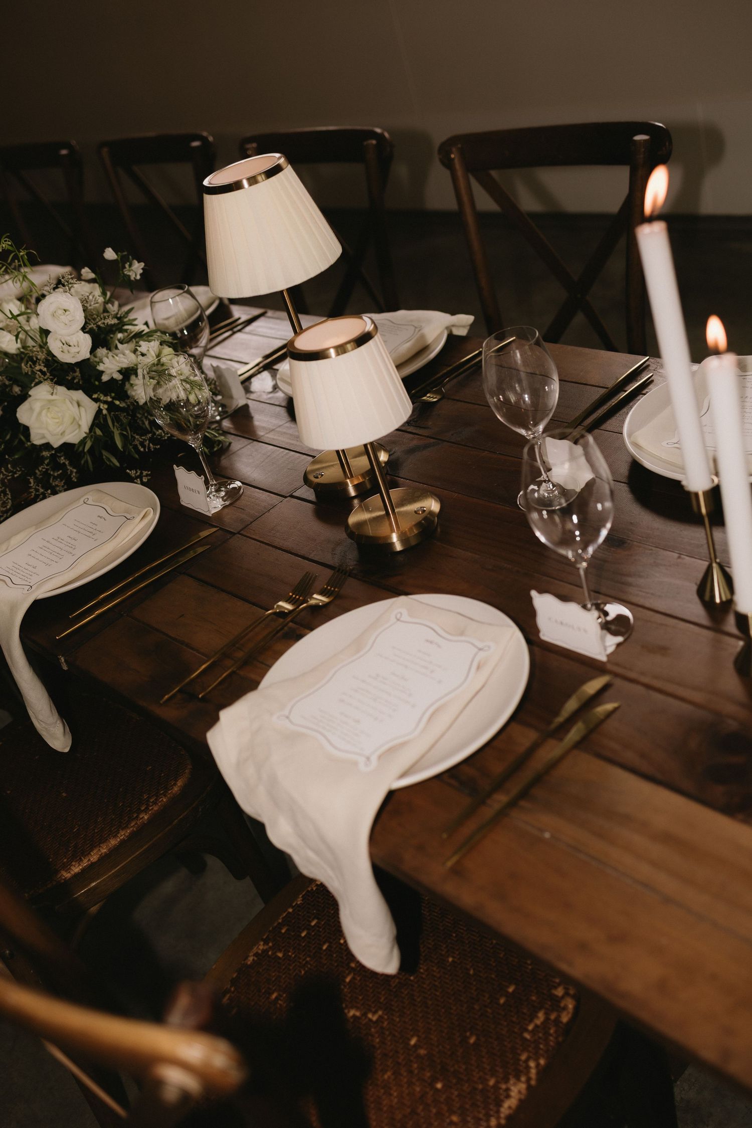 Elegant dark wood dining table with white candles, small brass lamps, crystal glasses, and white linen napkins for a wedding reception.