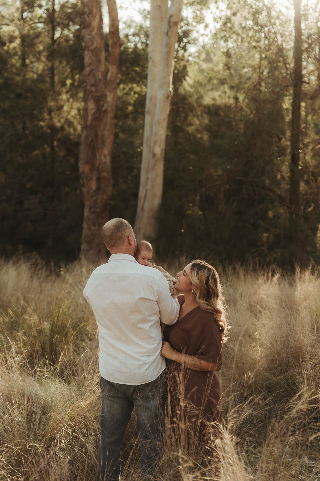 A couple stands in a sunlit field holding their baby, surrounded by tall golden grass and eucalyptus trees.