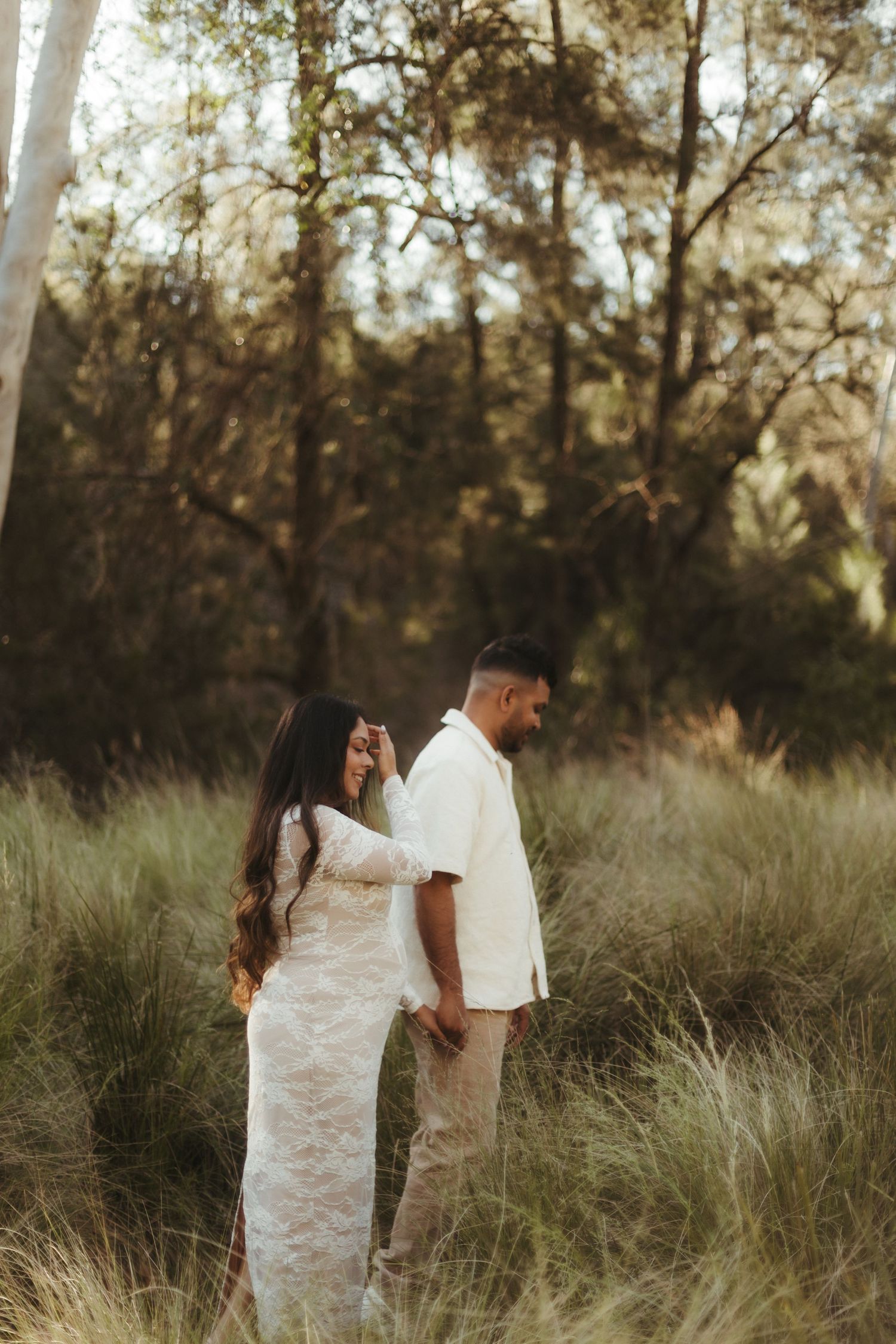 Expecting couple standing back to back in sunlit grassy woodland for maternity shoot.