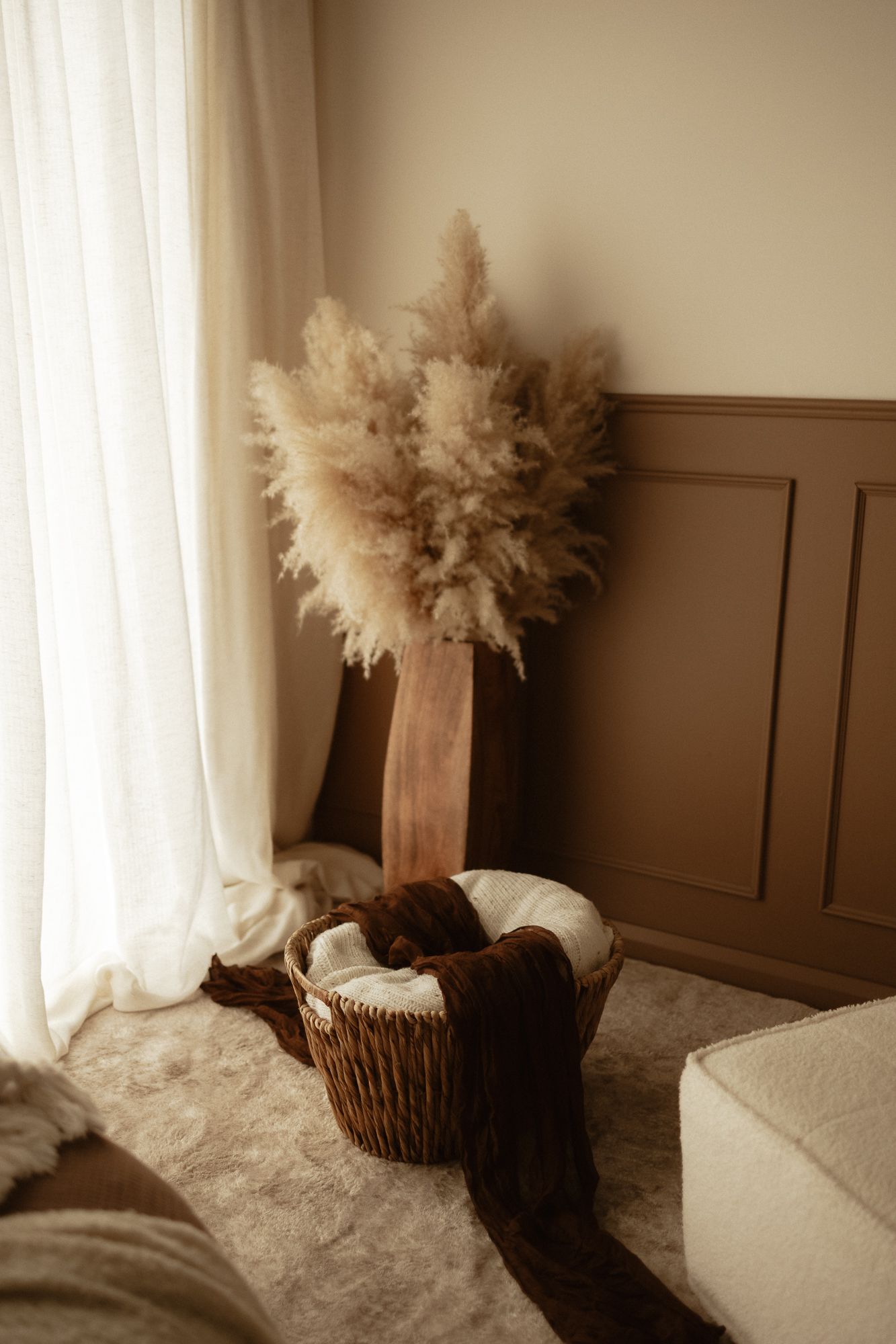 Boho interior corner with pampas grass in wooden vase, wicker basket with blanket, white curtains, and beige wainscoting.