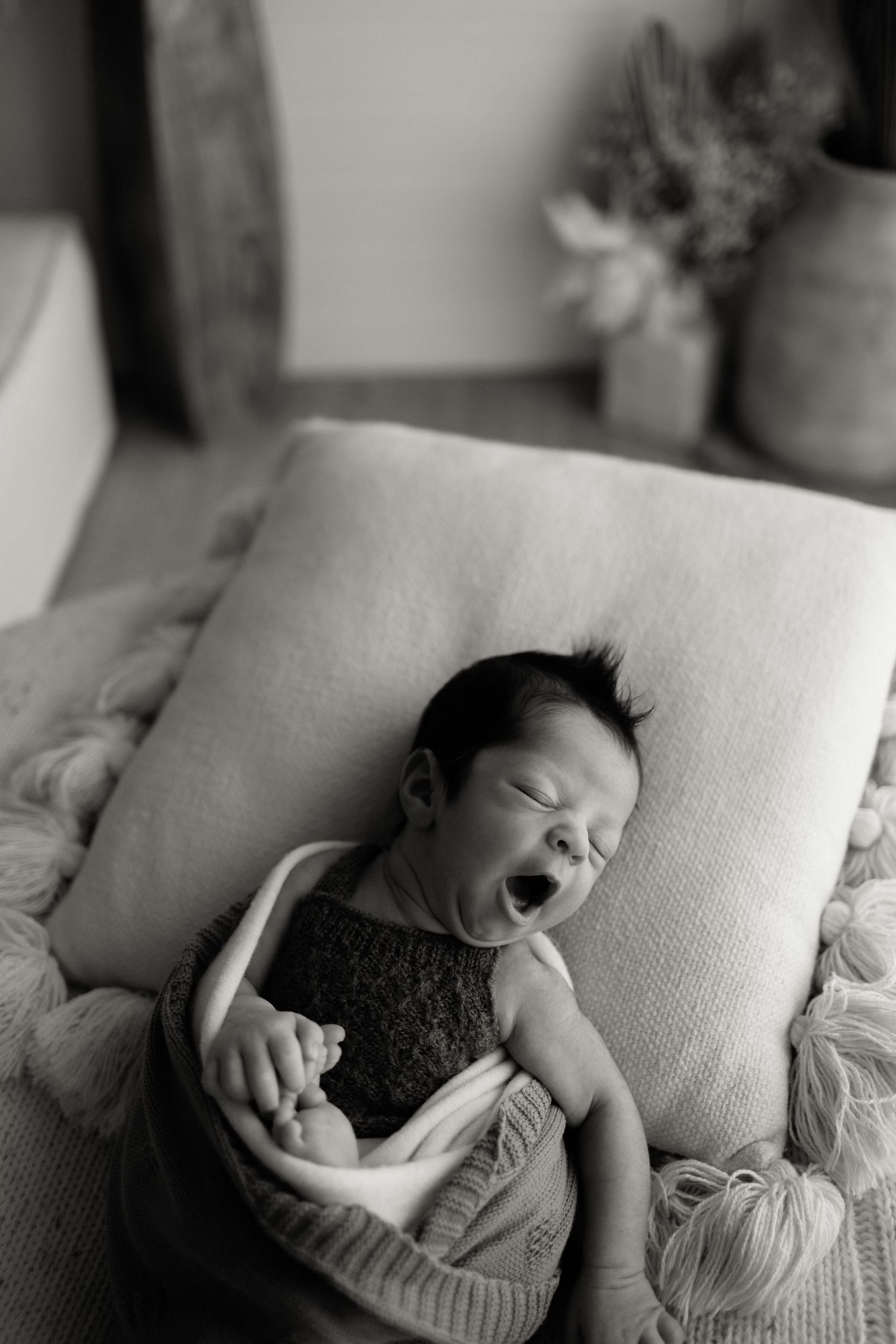 Black and white photo of a yawning newborn baby lying on a soft cushion and knit blanket.