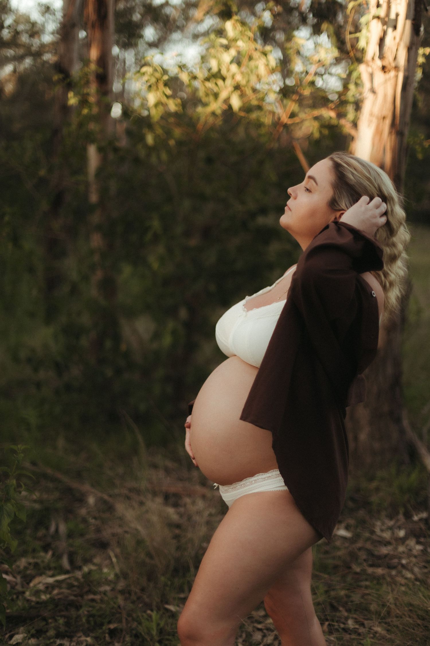 Pregnant woman in white lingerie and dark cardigan poses outdoors in golden forest light, cradling her baby bump.