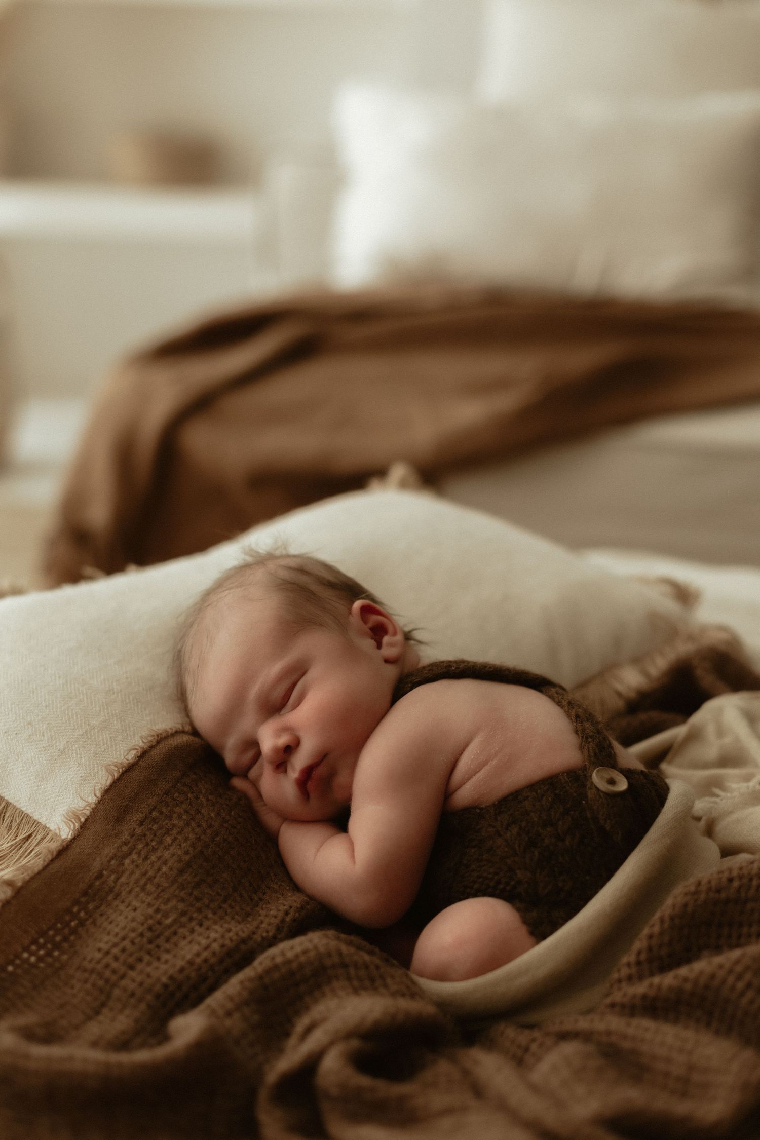 Sleeping newborn baby in brown knit romper resting peacefully on cream and brown blankets.