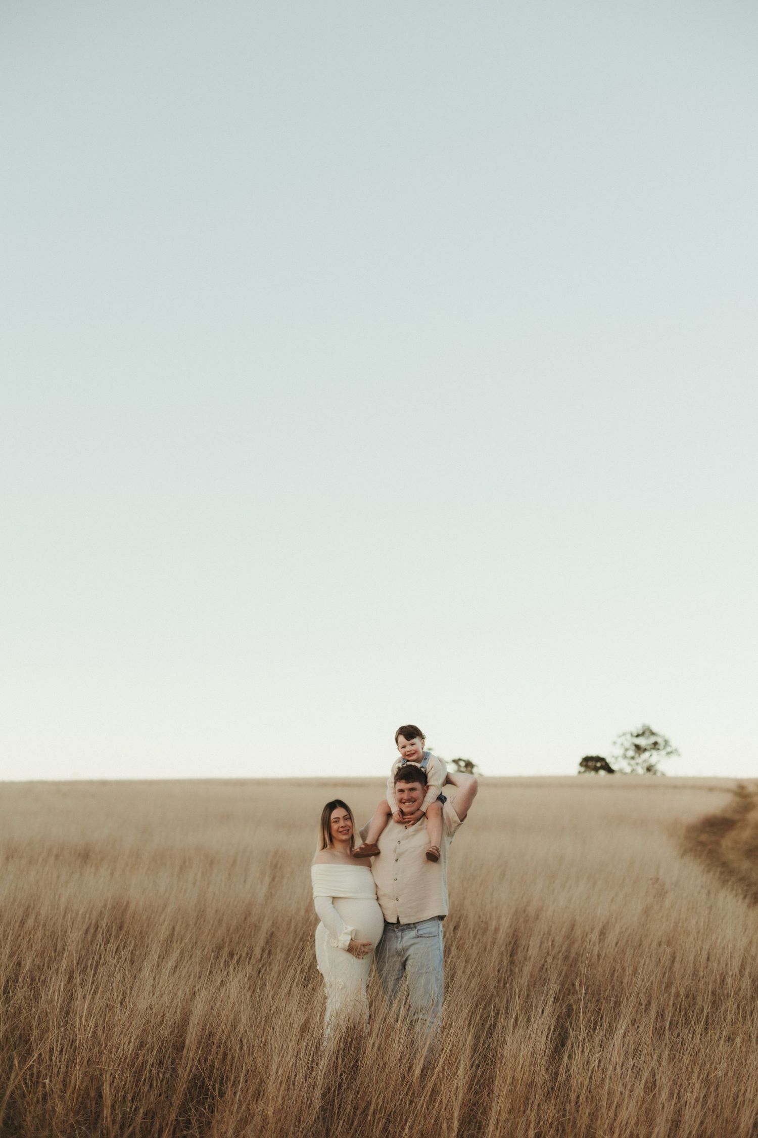 Pregnant woman and man holding toddler on shoulders standing in a vast golden grass field.