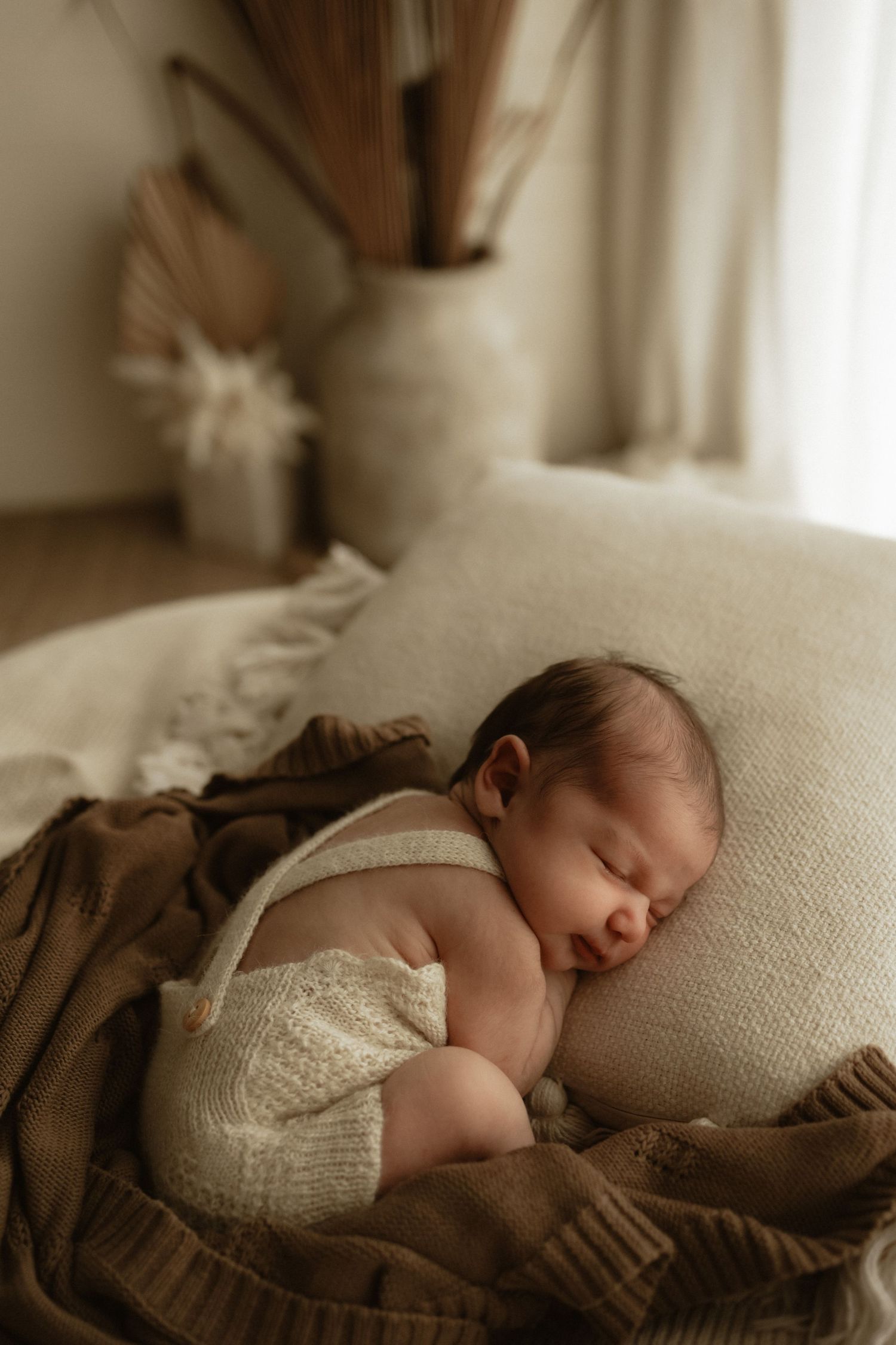 Sleeping newborn baby in cream knit overalls curled on a beige cushion with brown blankets in a warm, neutral nursery.