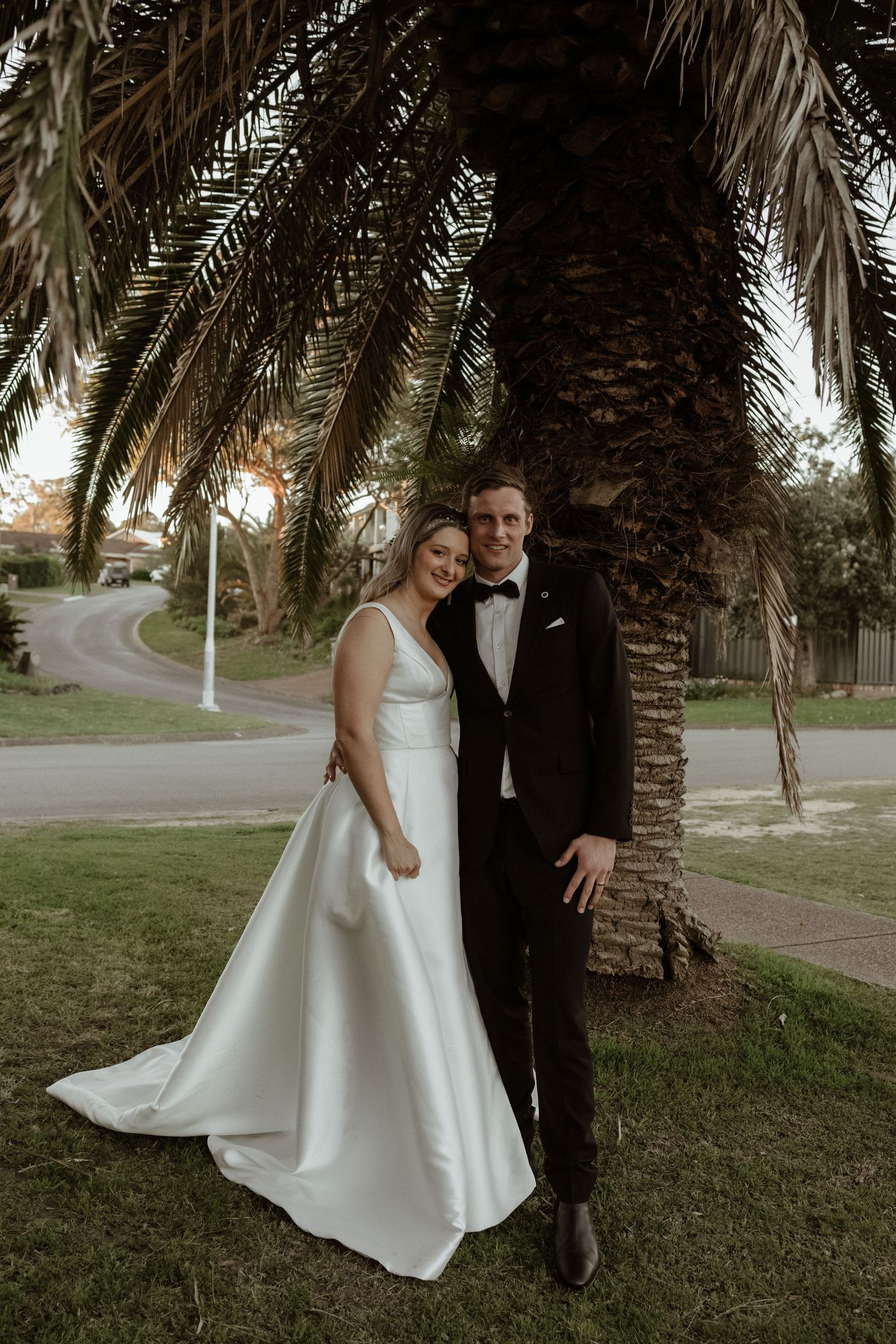 Bride in white gown and groom in black tuxedo posing together under a palm tree on their wedding day.